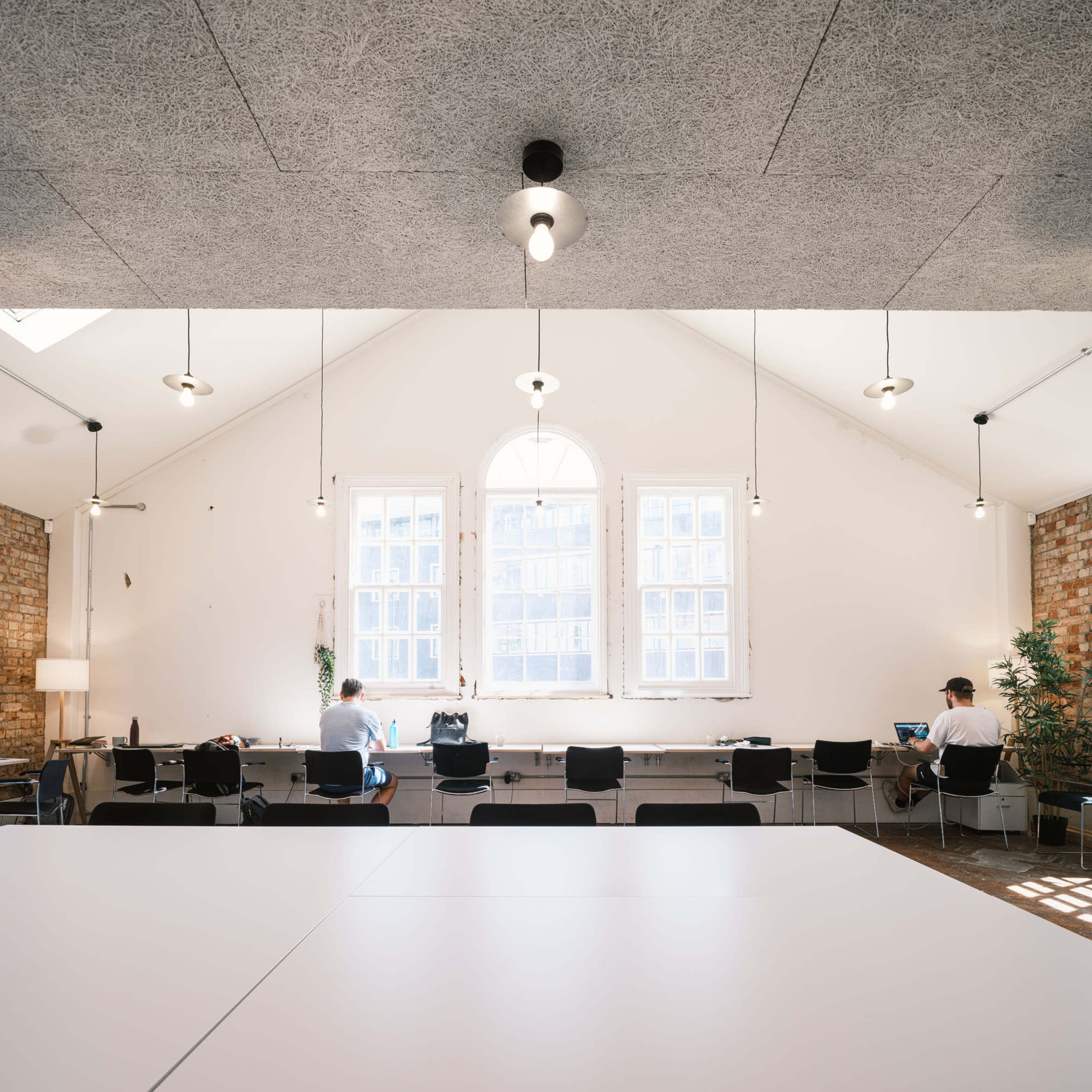 A spacious room with a vaulted ceiling, featuring long tables, hanging lights, and two individuals working at laptops near large windows.