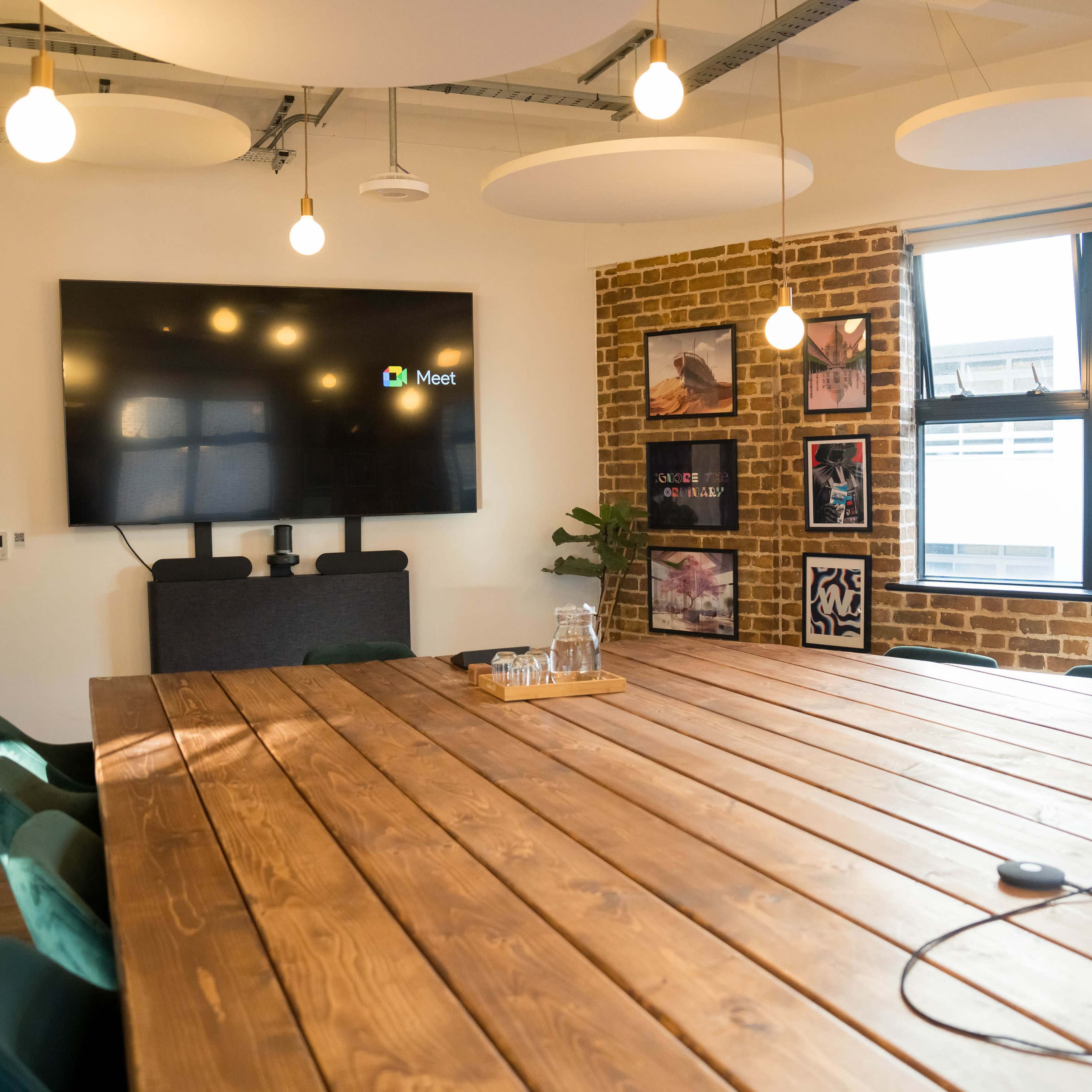 A large wooden conference table is surrounded by green chairs in a meeting room with a wall-mounted screen and exposed brick.