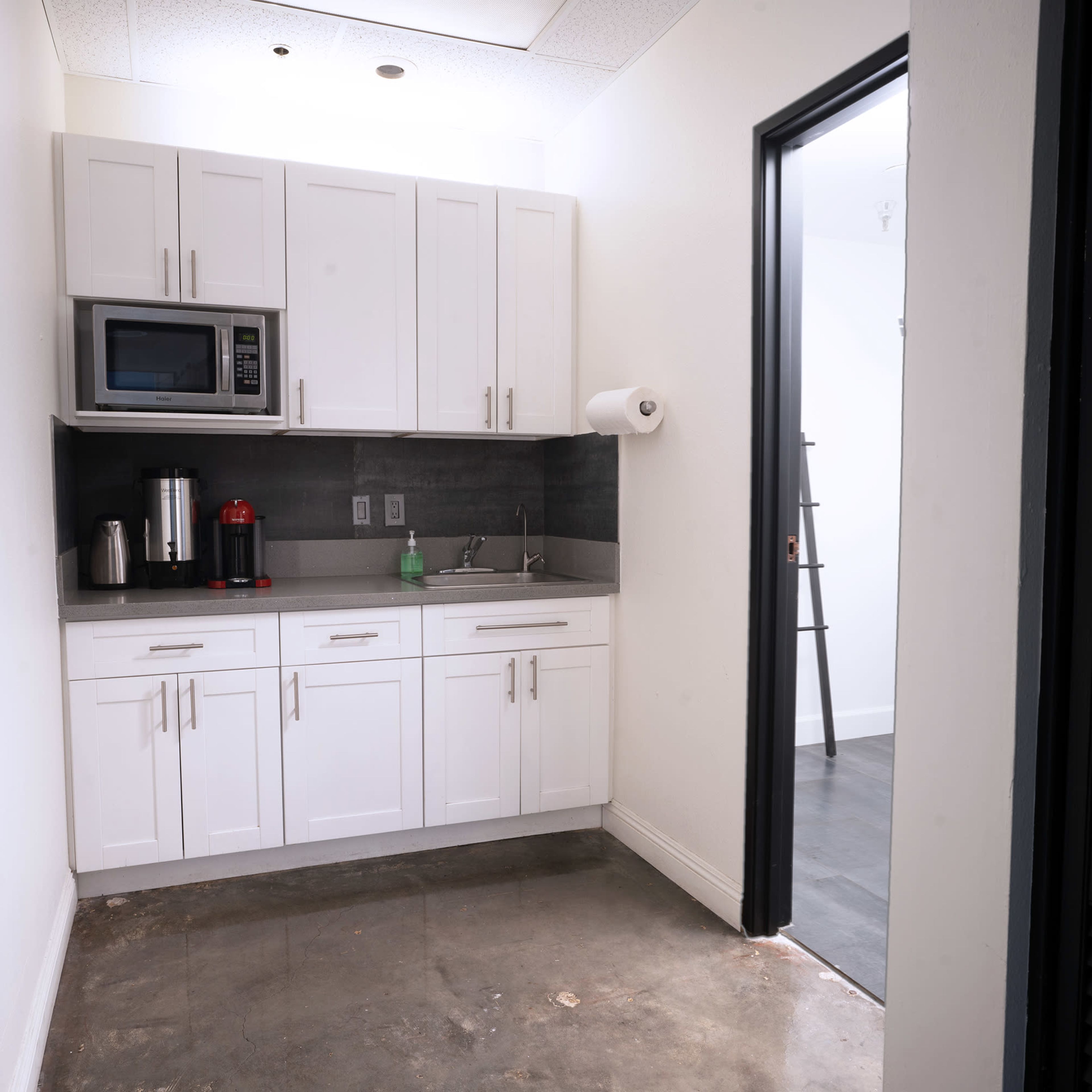 A small kitchen area with white cabinetry, a microwave, and a stainless steel sink, adjacent to a door leading to another room.