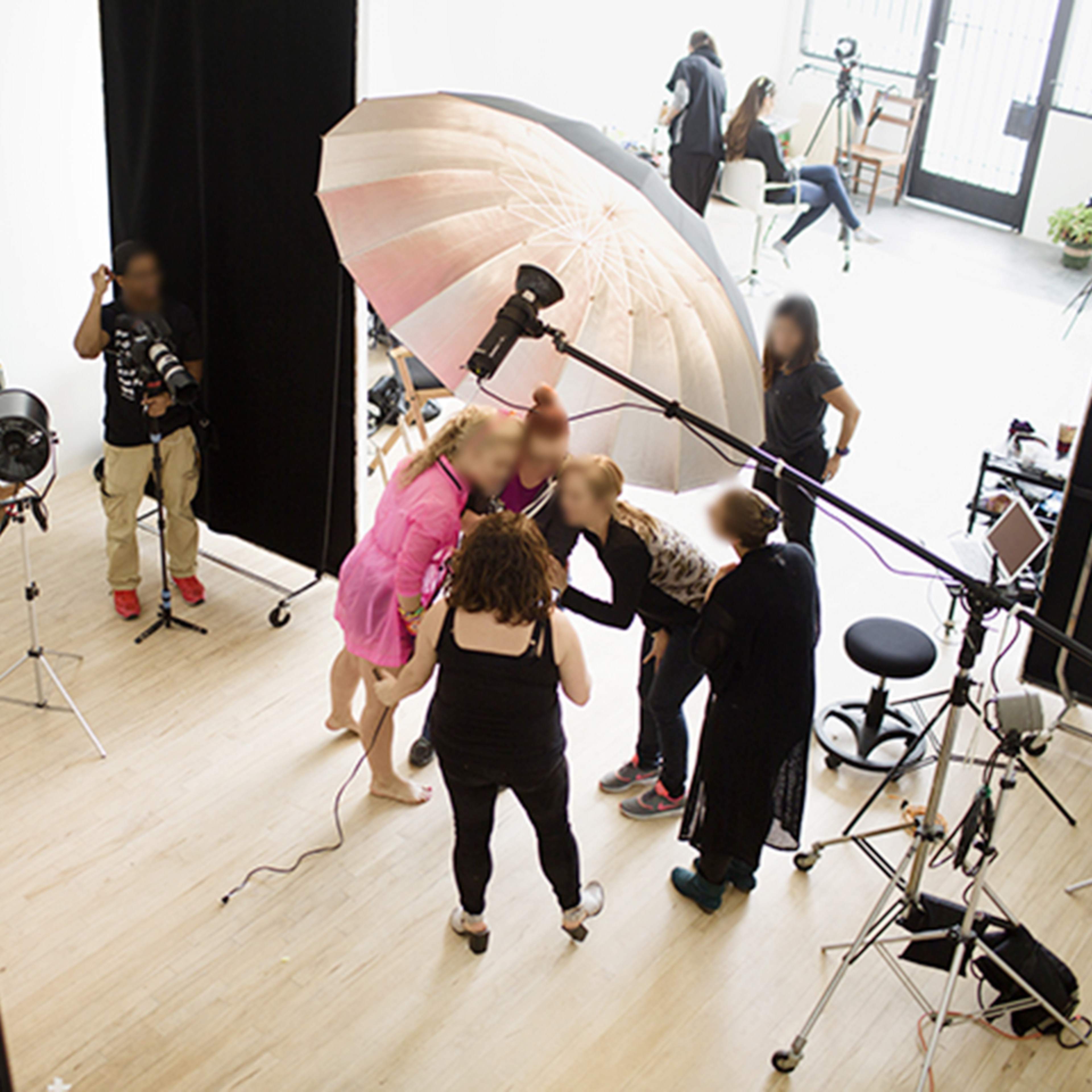 A photography studio setting features several people gathered around a subject, with a large umbrella light overhead and various equipment scattered throughout the space.