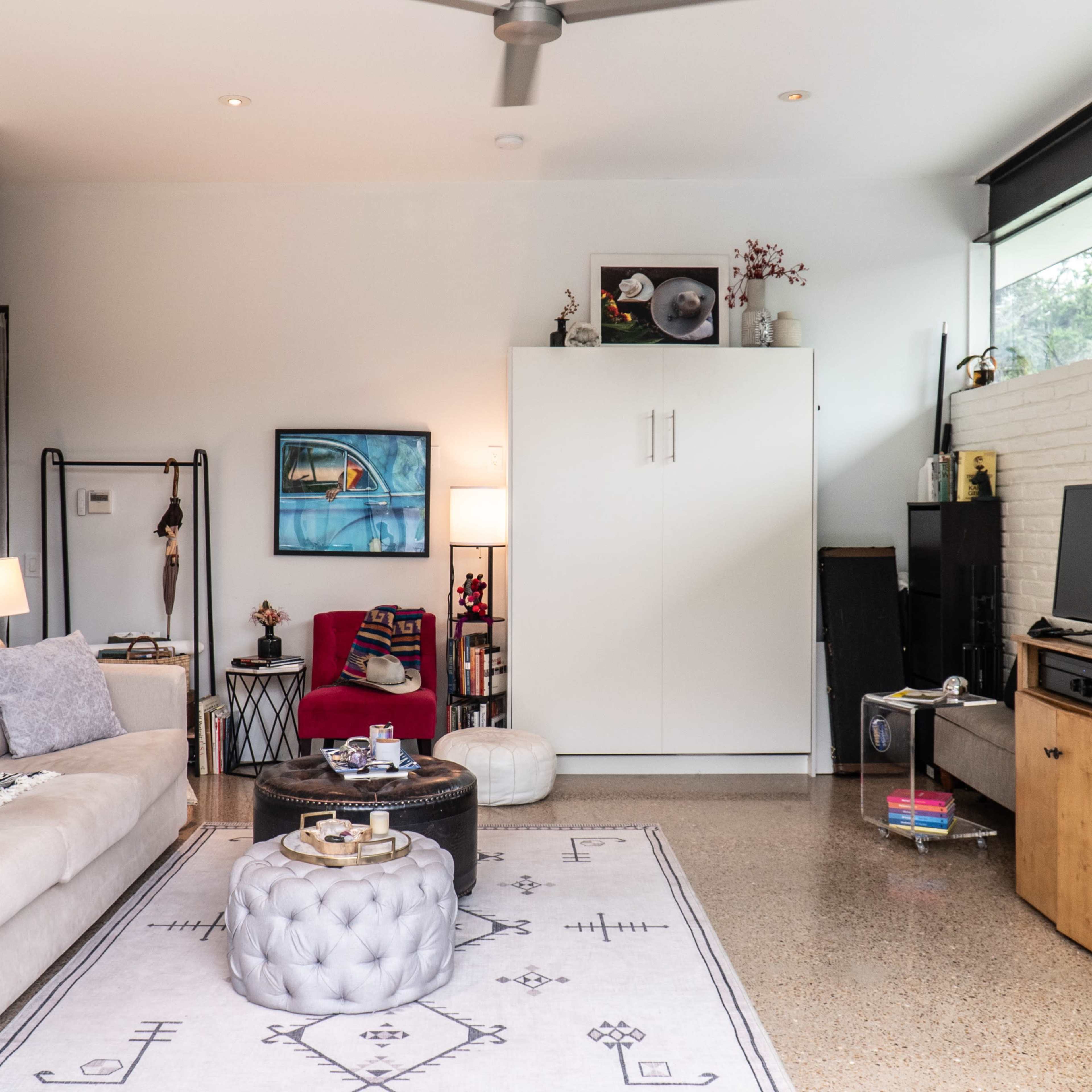 A light-filled living room features a beige sofa, a coffee table, and various shelves with decorations and a television against a white brick wall.