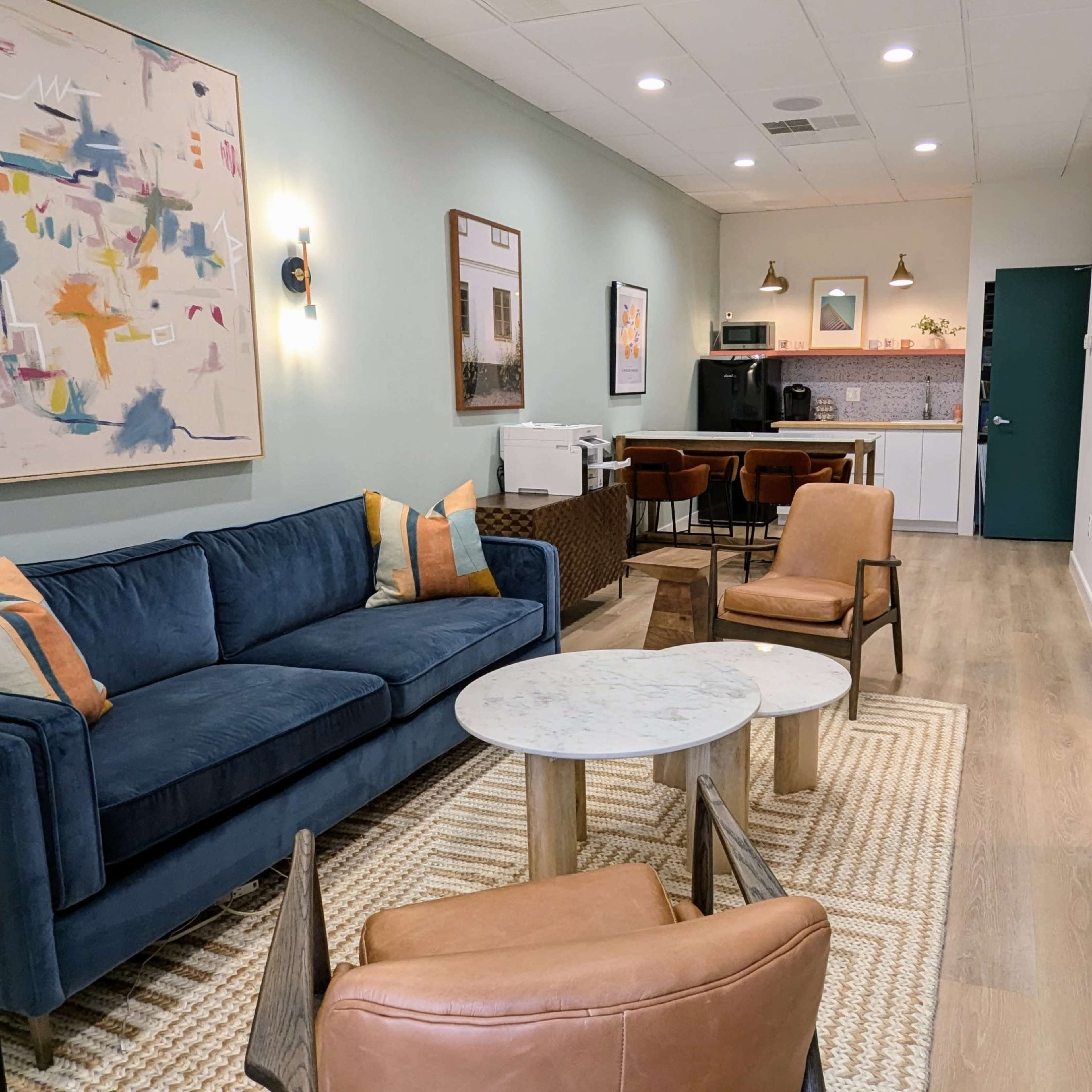 A well-lit, modern living space featuring a blue sofa, brown armchairs, a marble coffee table, and a kitchenette in the background.