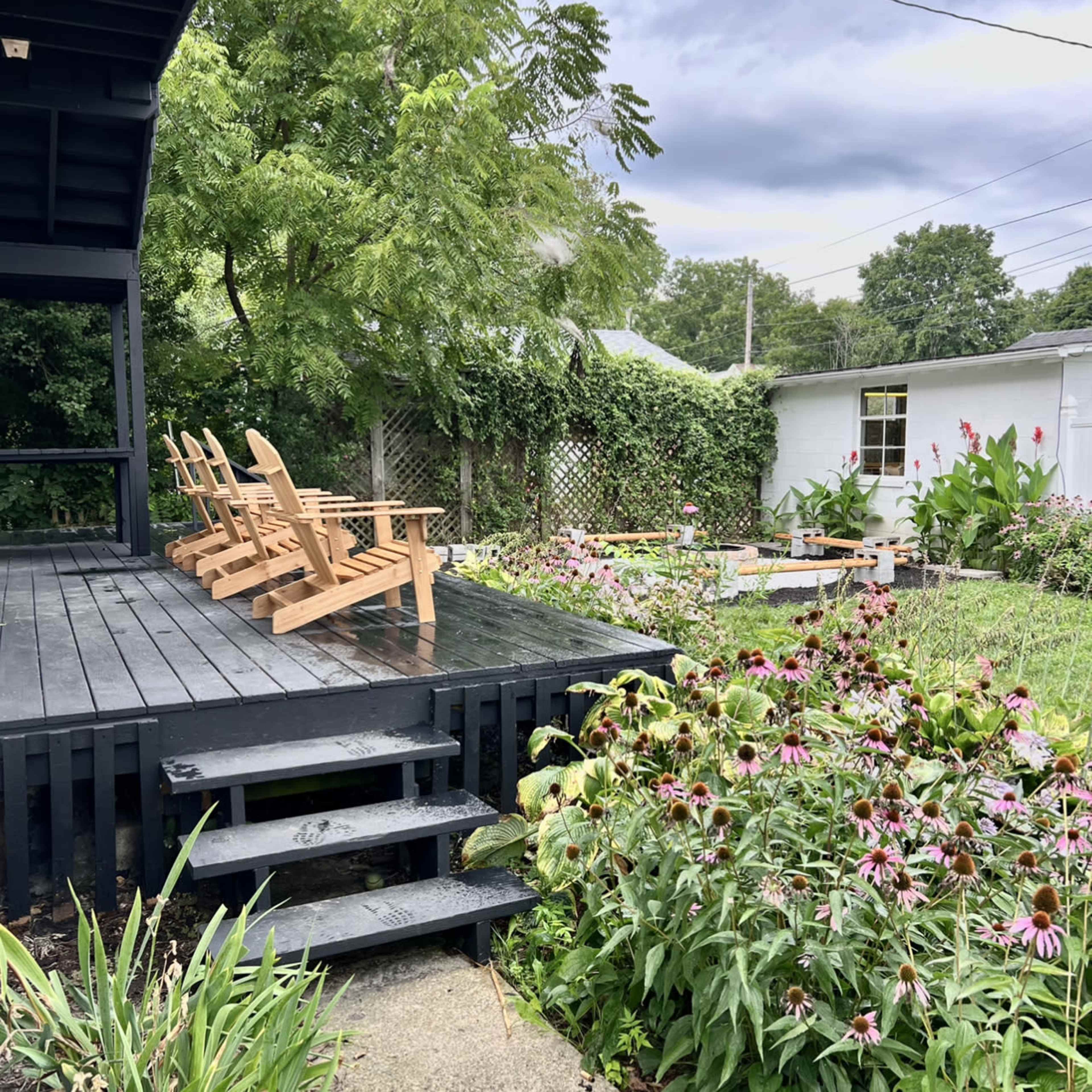 A wooden deck with several Adirondack chairs overlooks a garden filled with blooming flowers and greenery.