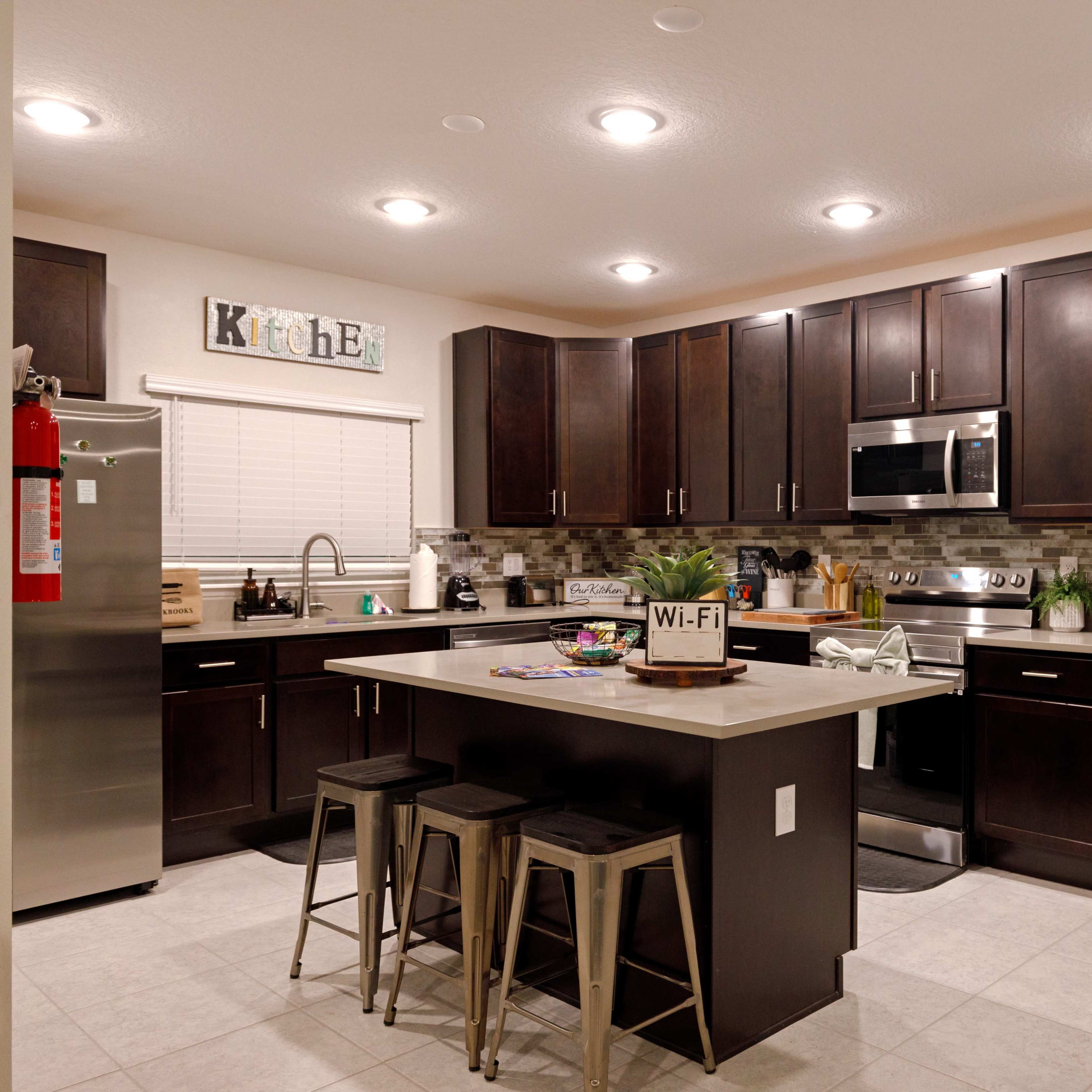 A modern kitchen with dark wood cabinets, stainless steel appliances, and a central island with three metal bar stools.