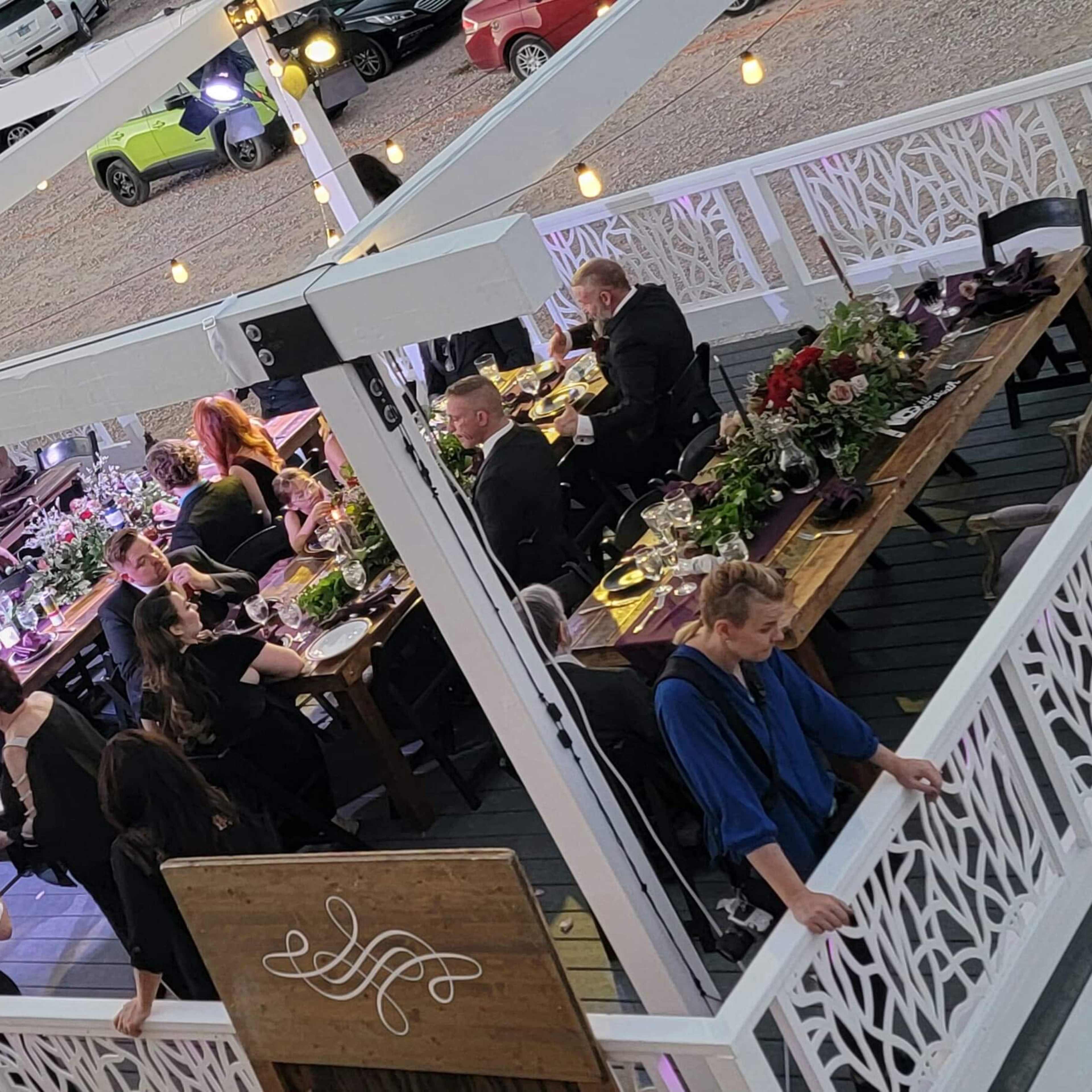 A dining setup at an outdoor venue, with guests seated around tables adorned with flowers, surrounded by desert scenery and vehicles in the background.