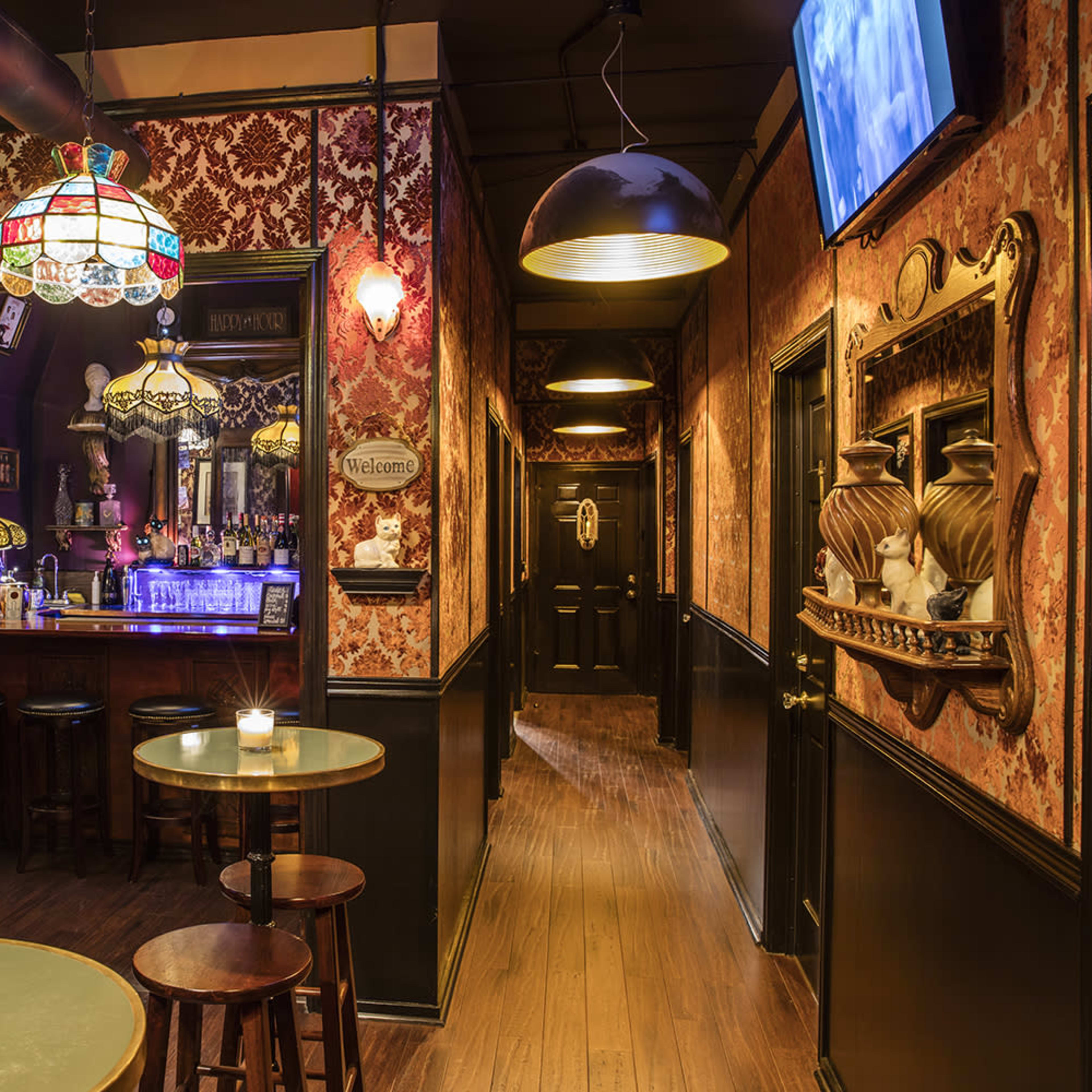 A dimly lit bar with a long hallway adorned with vintage decor, leading to a bar area featuring a stained glass lamp and several seating tables.