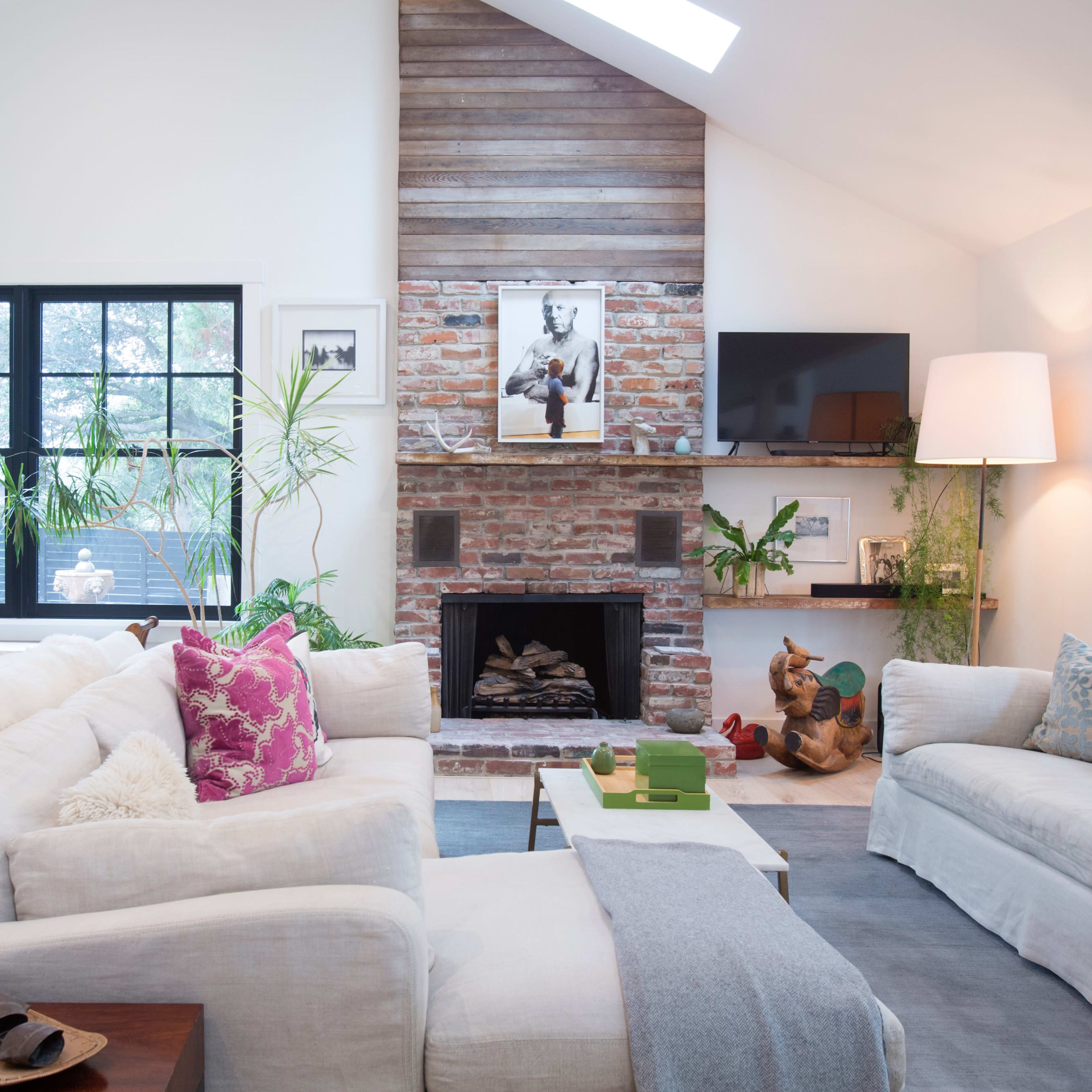 A modern living room with two large sofas, a brick fireplace, plants, and large windows allowing natural light.