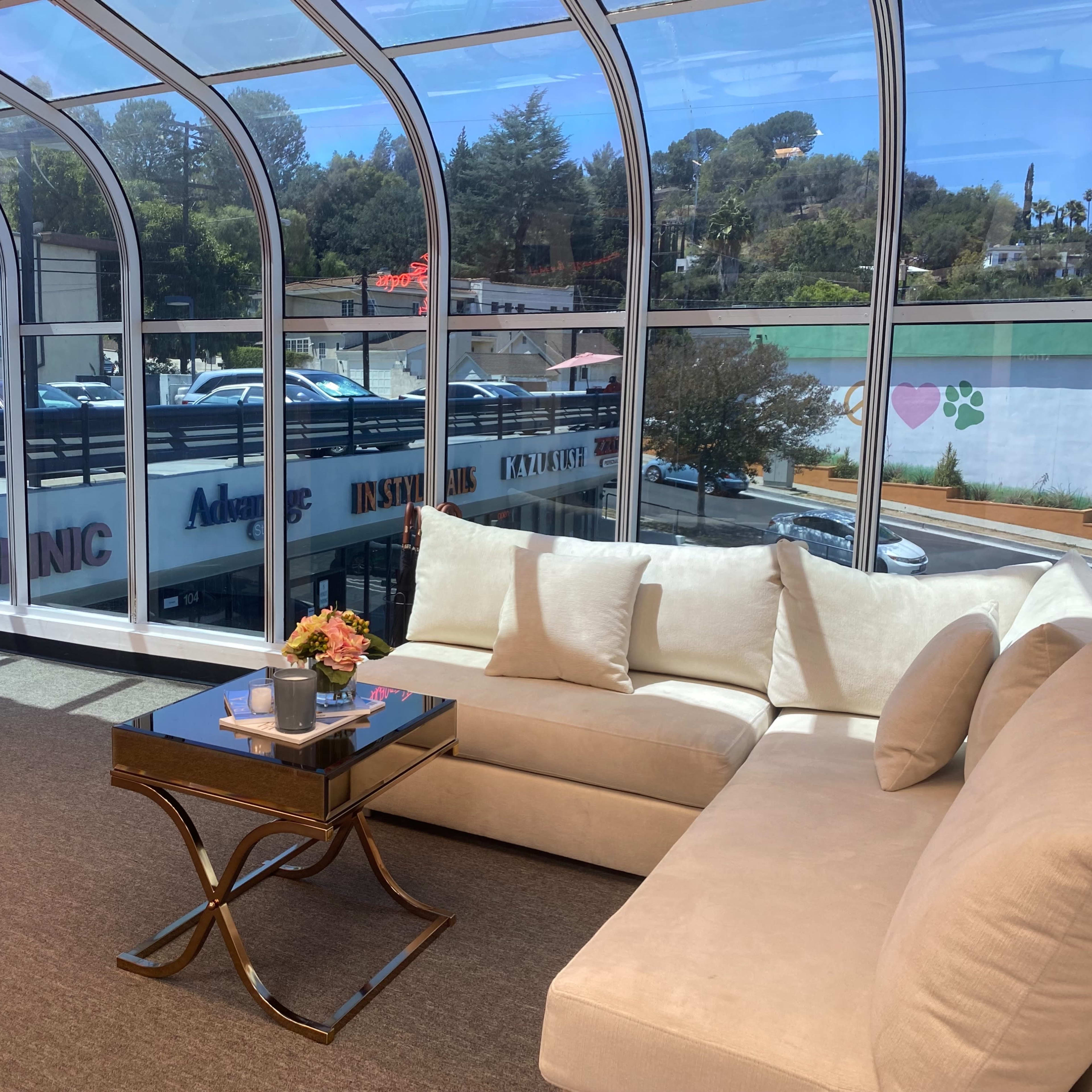 A modern seating area with a large light-colored sofa, a small coffee table, and large windows overlooking a parking lot and nearby buildings.