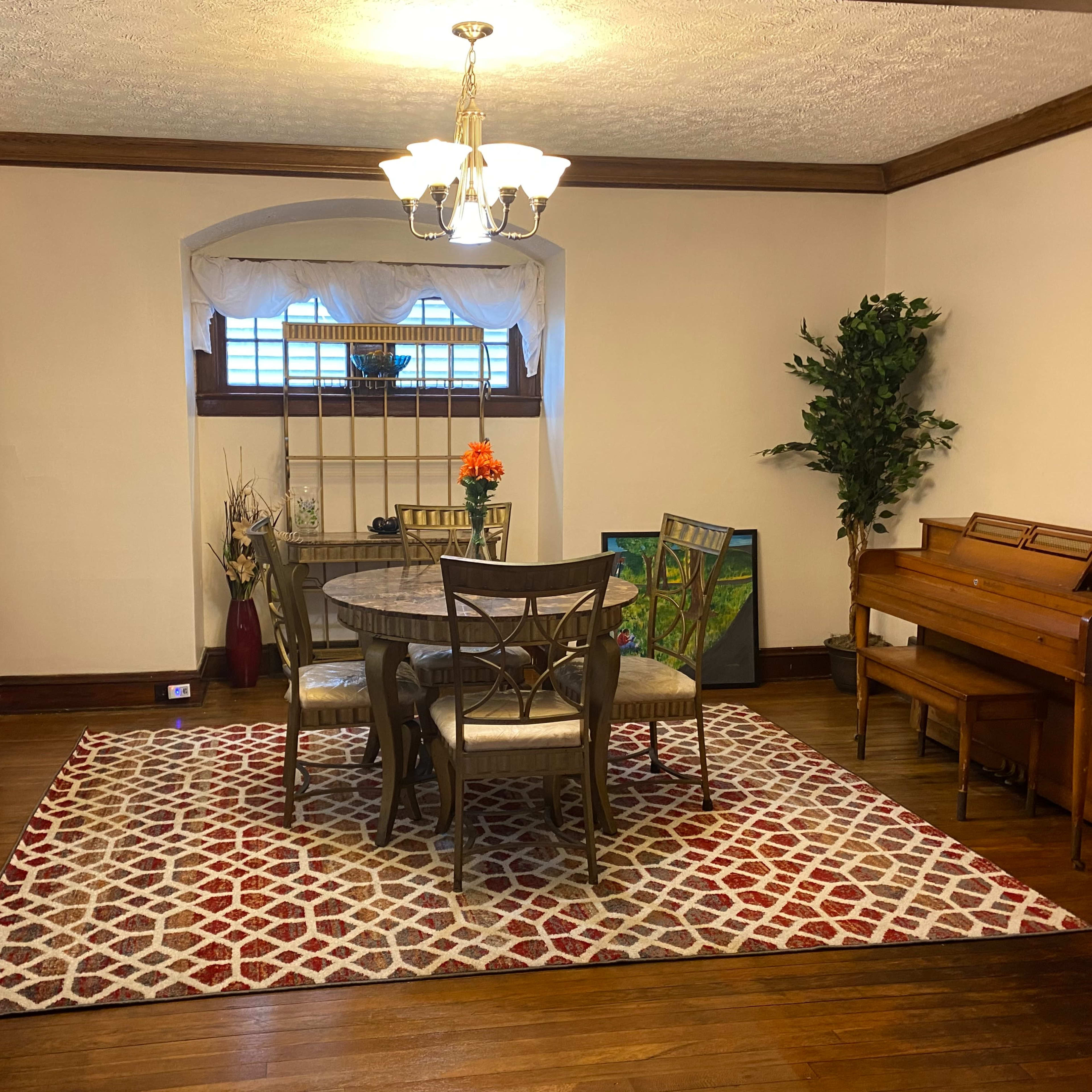 A dining area with a round table and four chairs, surrounded by a patterned rug, a wooden piano, and a display cabinet.