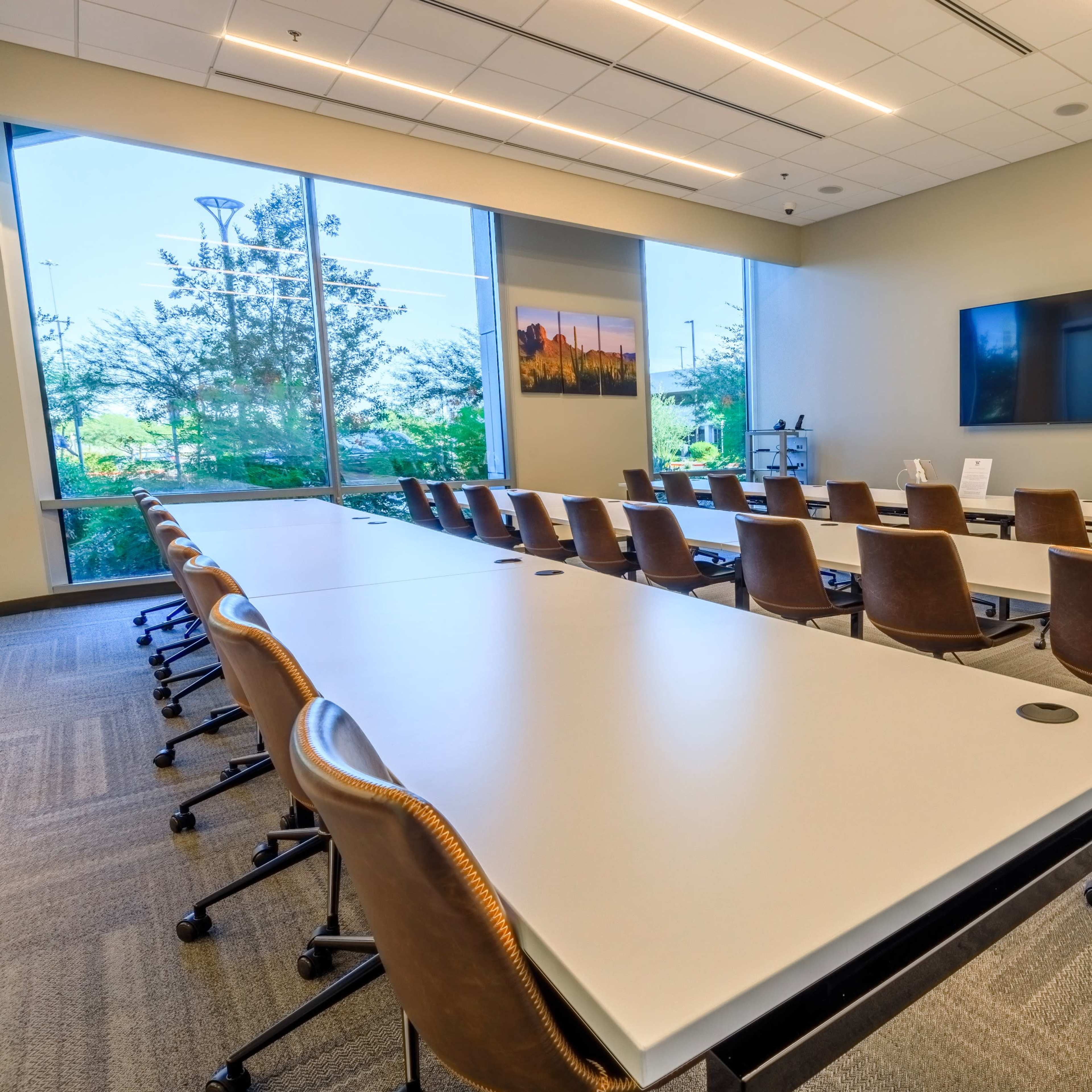 A modern conference room features rows of tables and chairs facing a large screen, with natural light streaming in through a window.