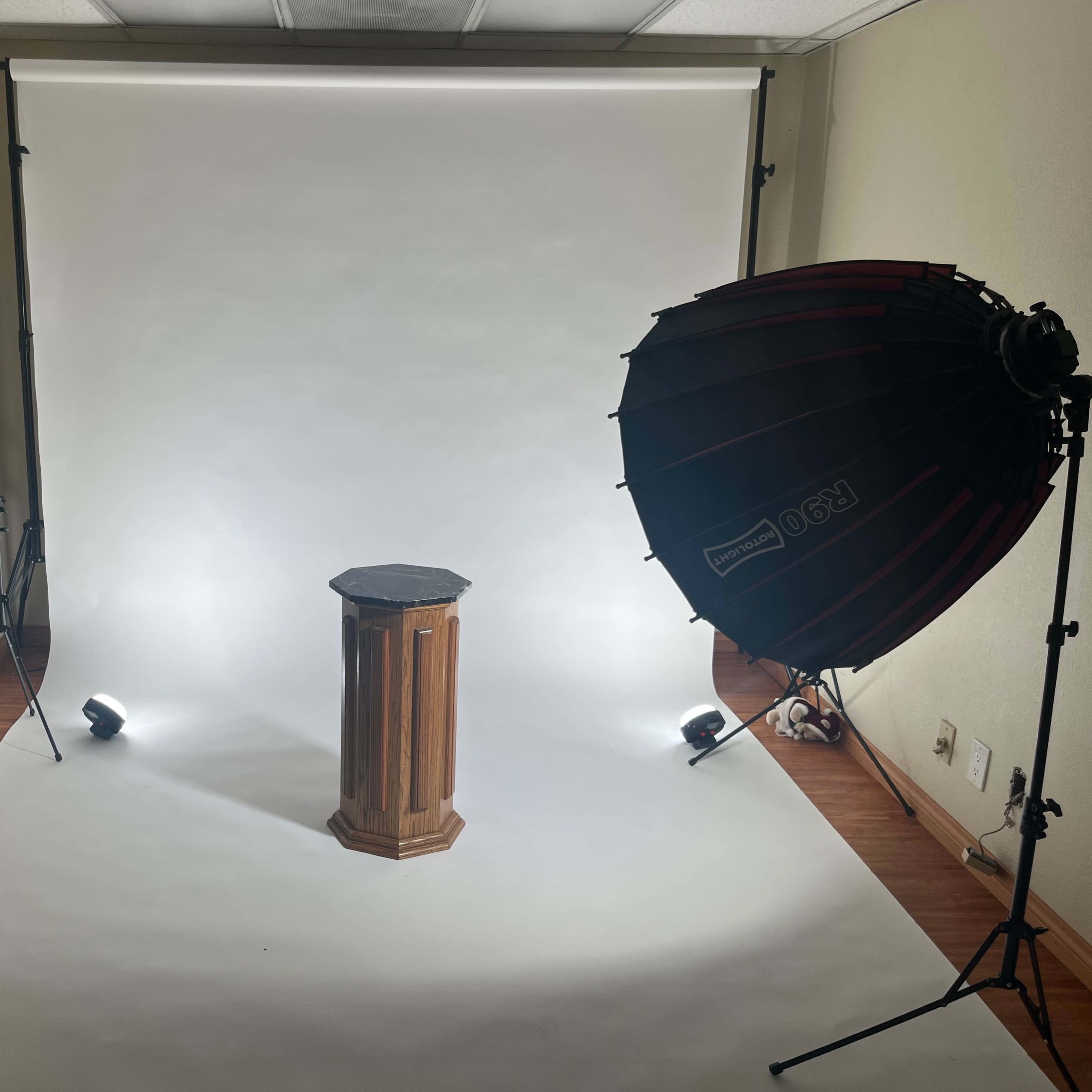 A photography studio with a wooden pedestal in the center, surrounded by lighting equipment and a white backdrop.