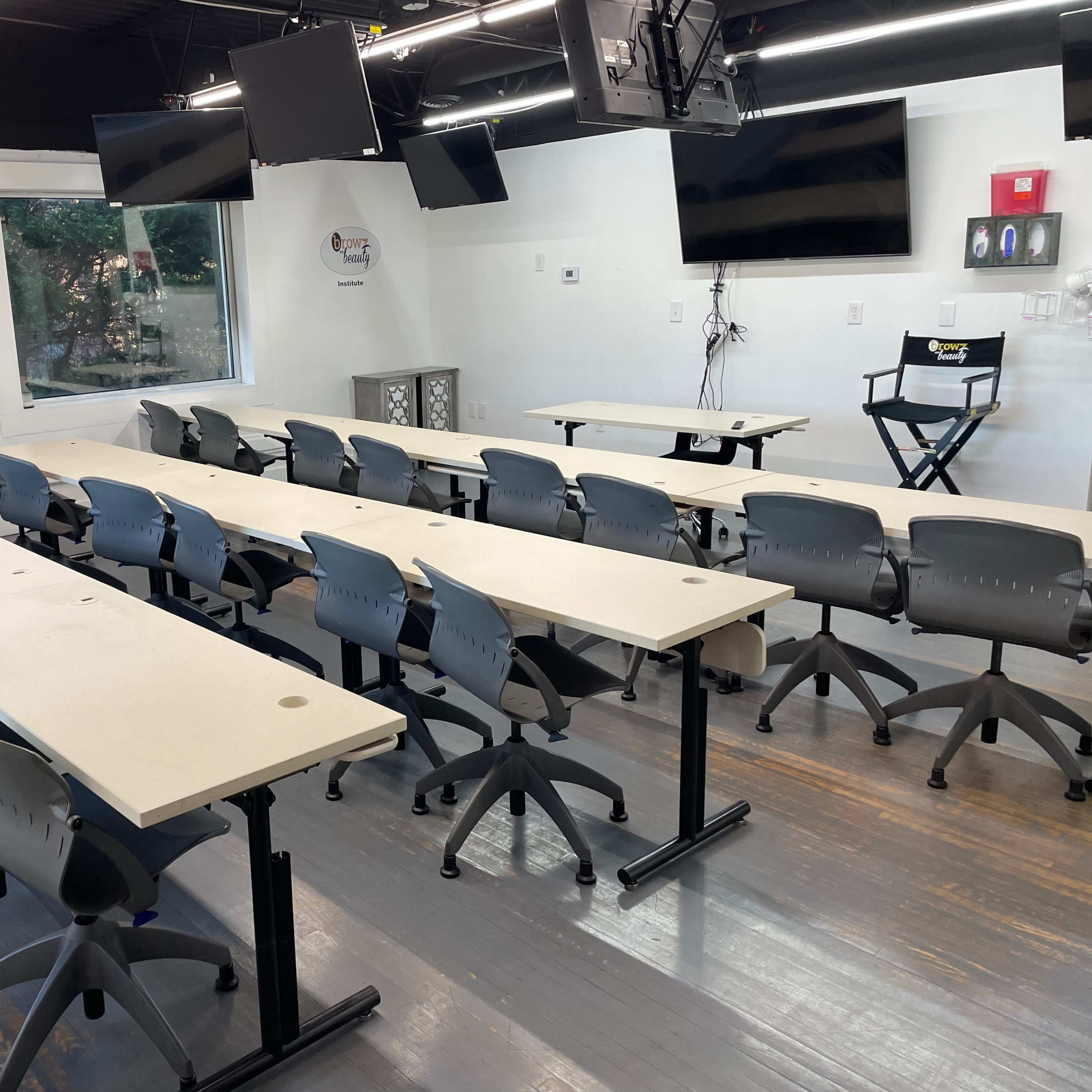 A spacious classroom setting with multiple rows of long tables and rolling chairs, equipped with monitors on the walls.