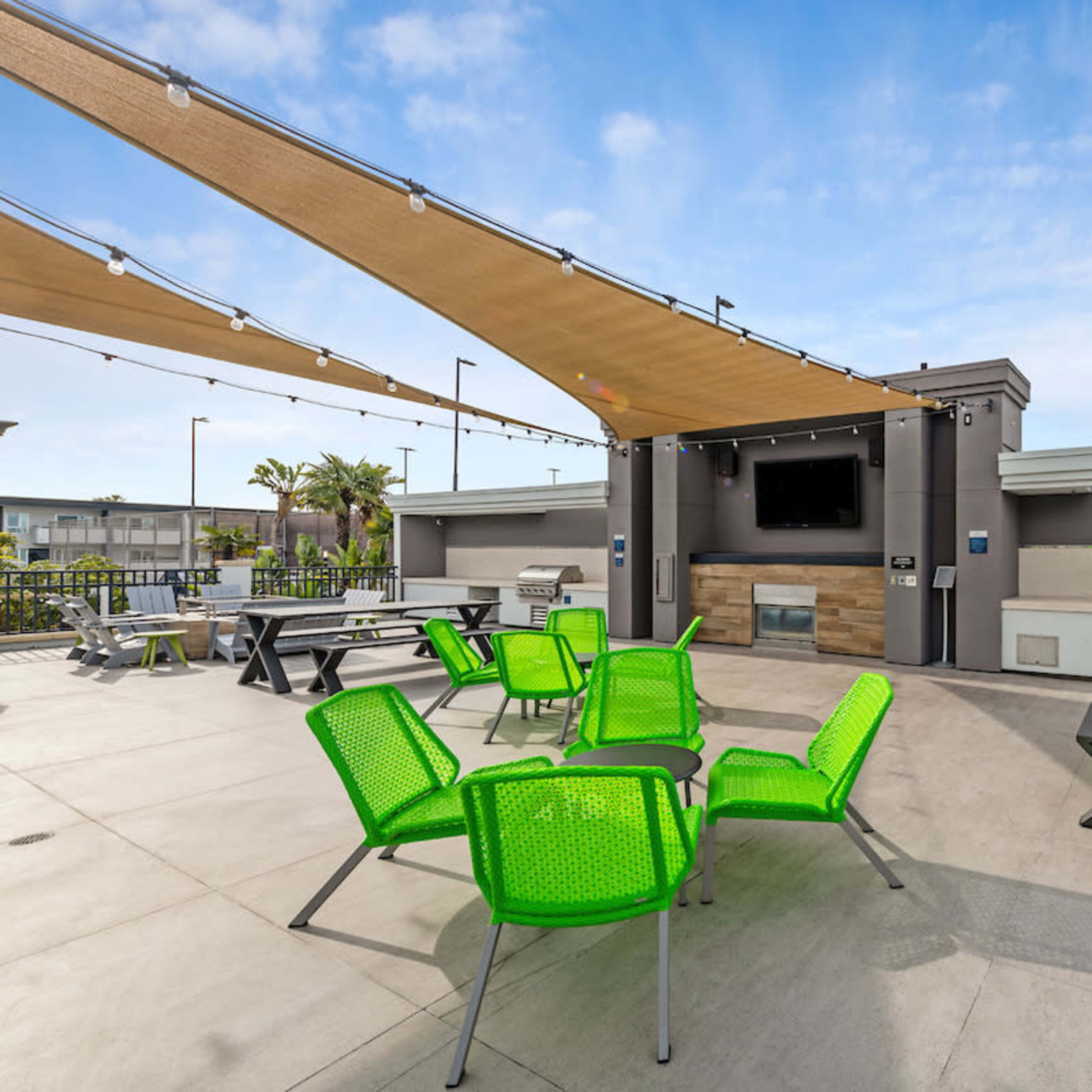 A rooftop terrace area equipped with green chairs, picnic tables, a grill, and a large screen.