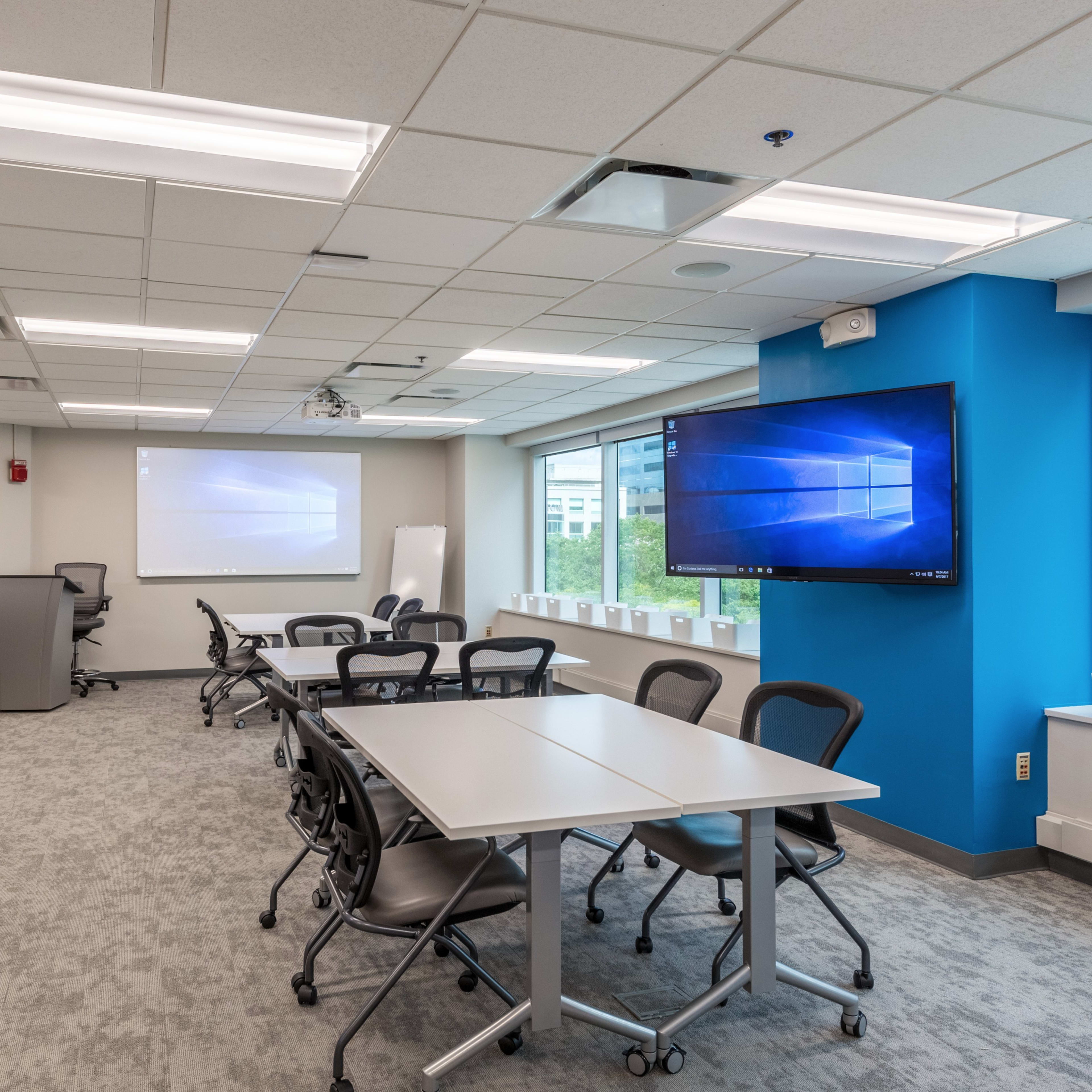 A modern conference room features a large screen, multiple tables, and ergonomic chairs, with natural light coming through the windows.