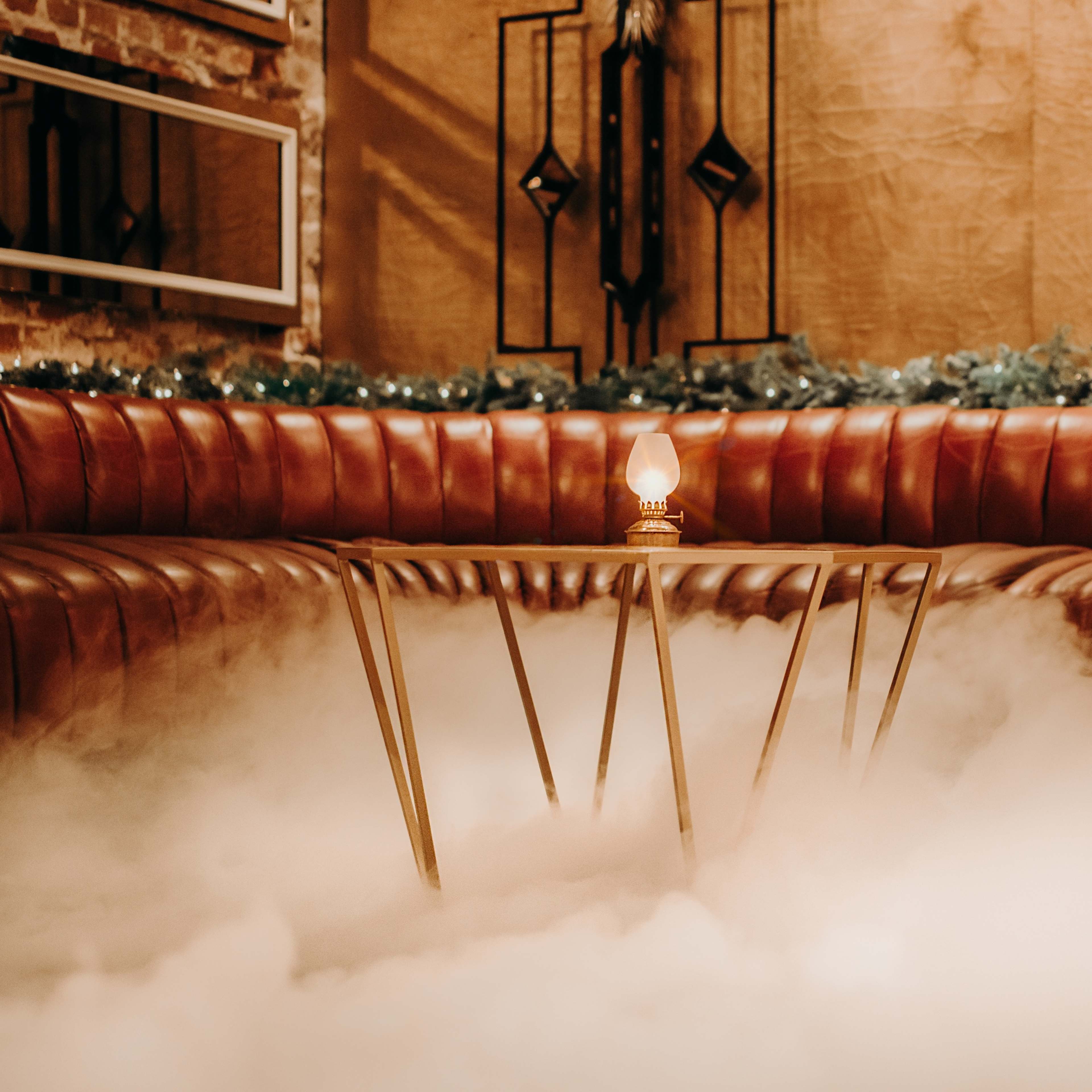 A low table with a lamp sits on a fog-covered floor in a dimly lit, cushioned seating area.