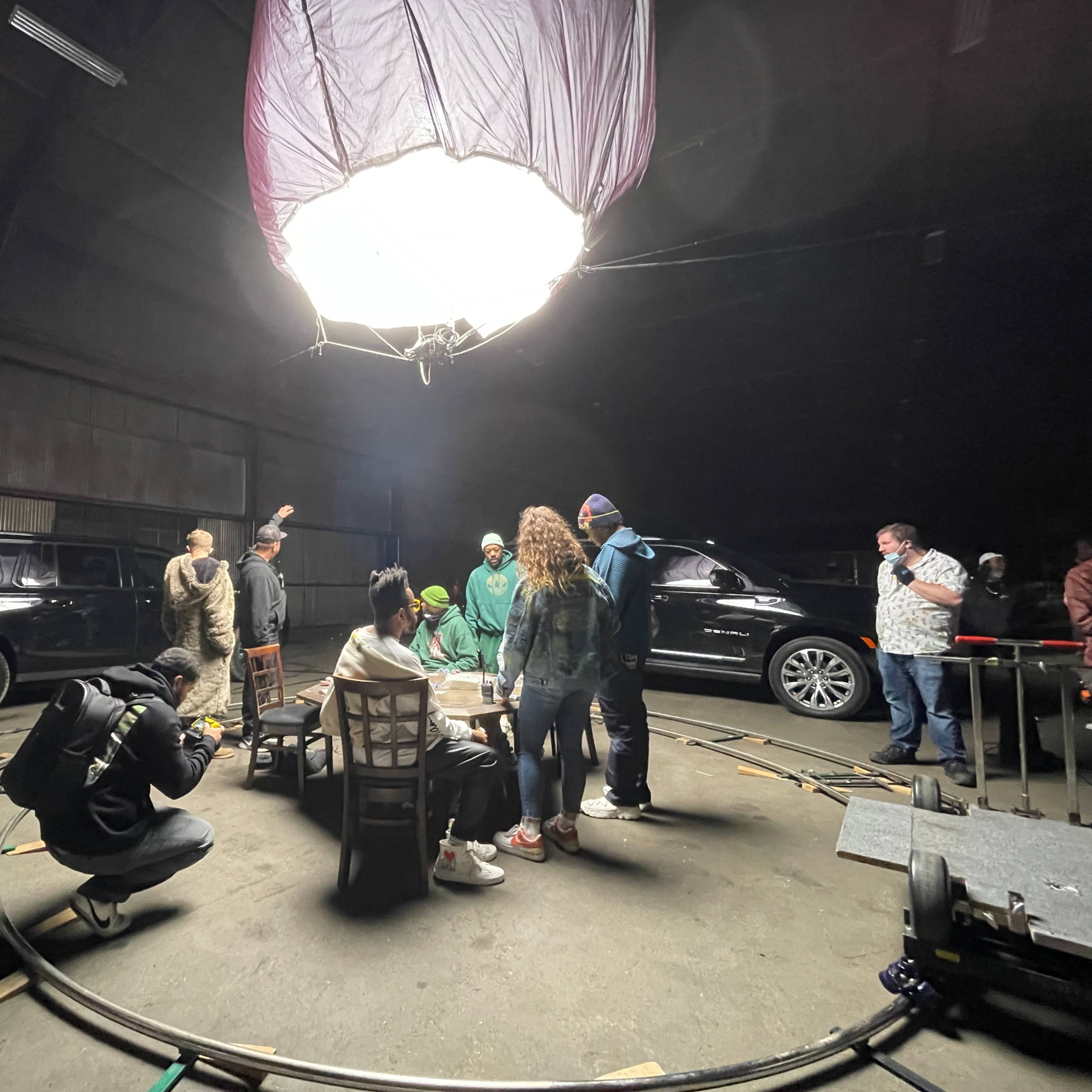 A group of people is gathered in a dimly lit warehouse, with film equipment and vehicles surrounding them during a shoot.