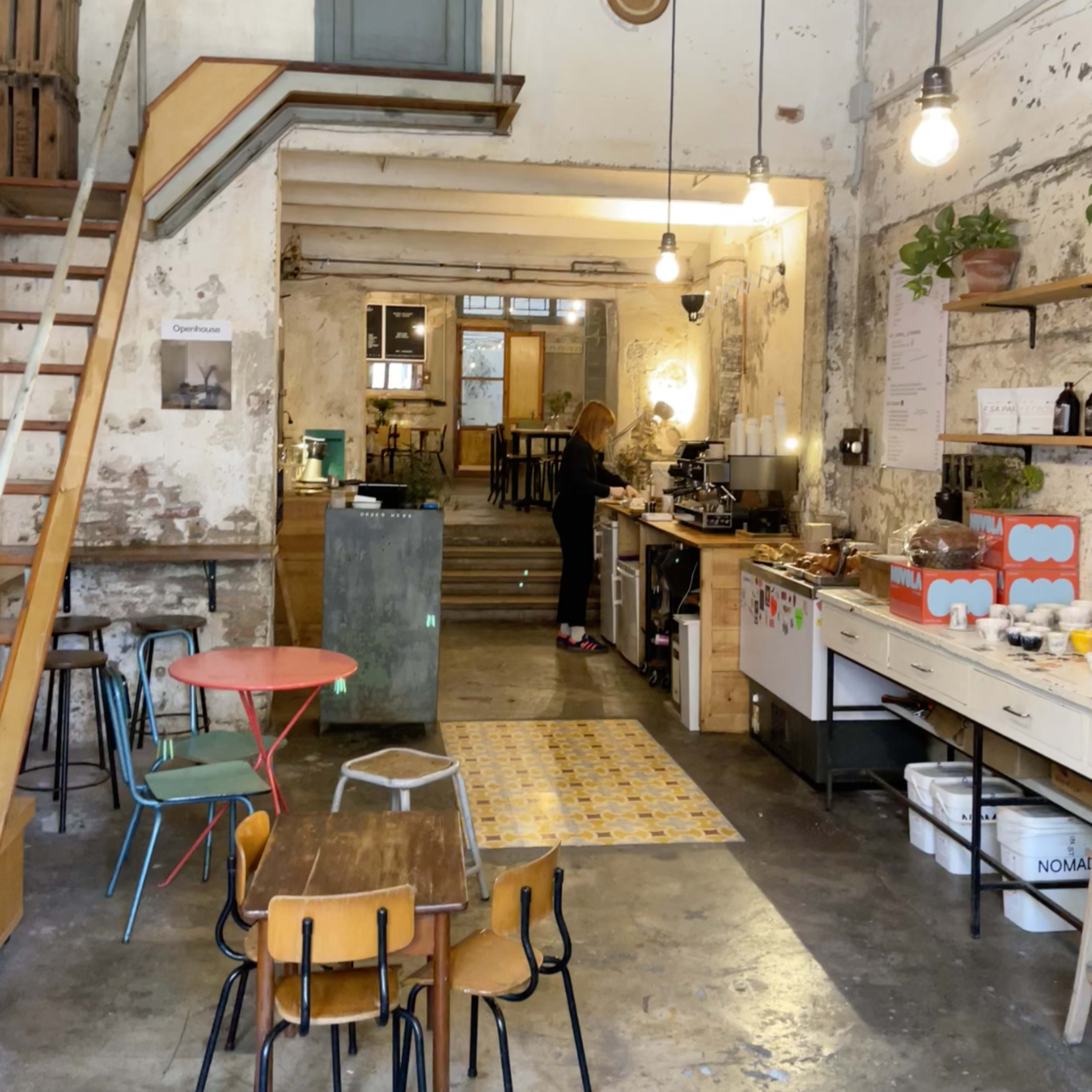 A spacious, rustic café with a staircase on the left, a woman preparing coffee at the counter, and various seating arrangements scattered throughout the room.