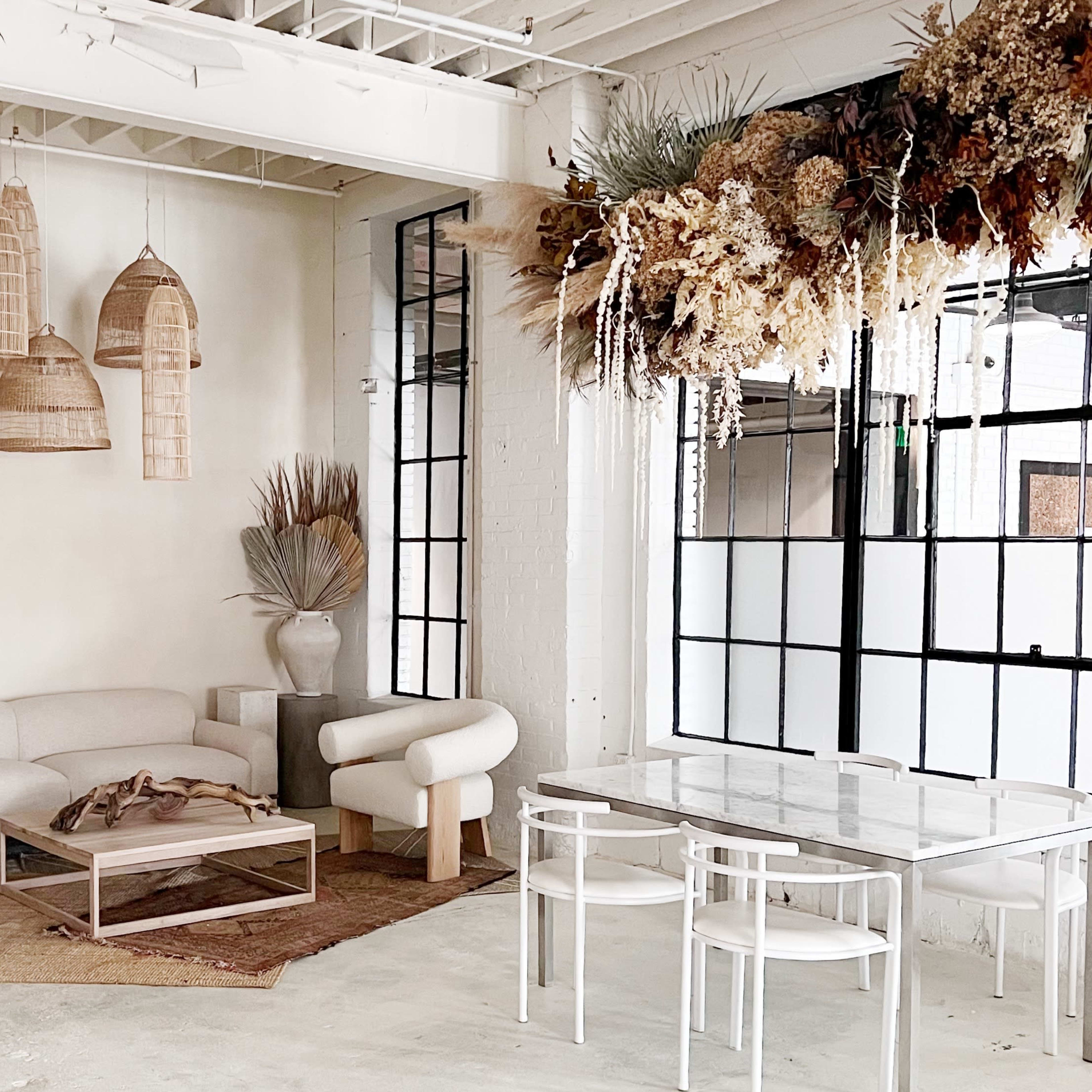A minimalist interior with a light-colored sofa, a wooden coffee table, and a dining area with transparent chairs, surrounded by decorative hanging baskets and dried floral arrangements.