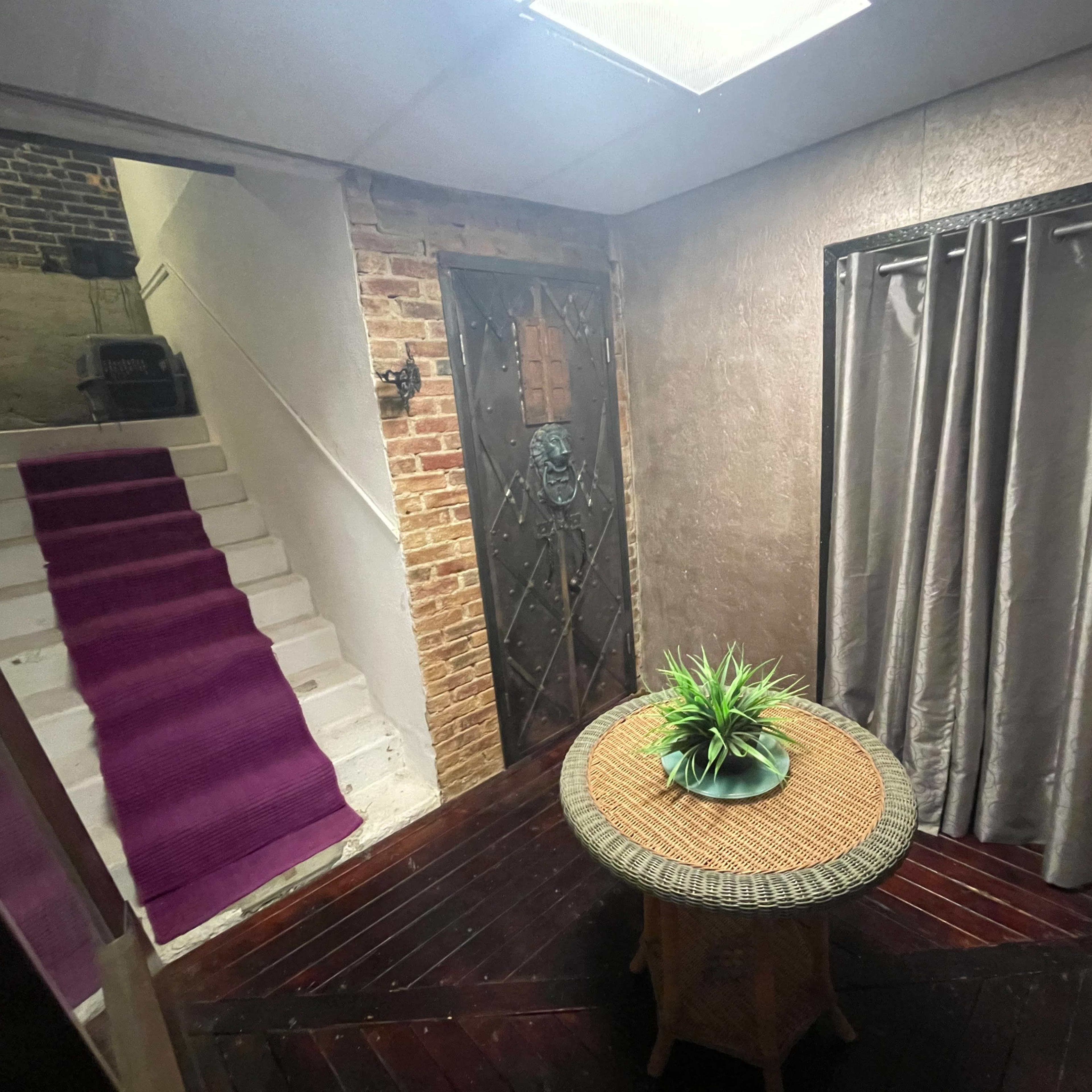 An entrance hall featuring a round table with a plant, a large door with a lion's head knocker, and a staircase with a purple runner leading to an upper level.