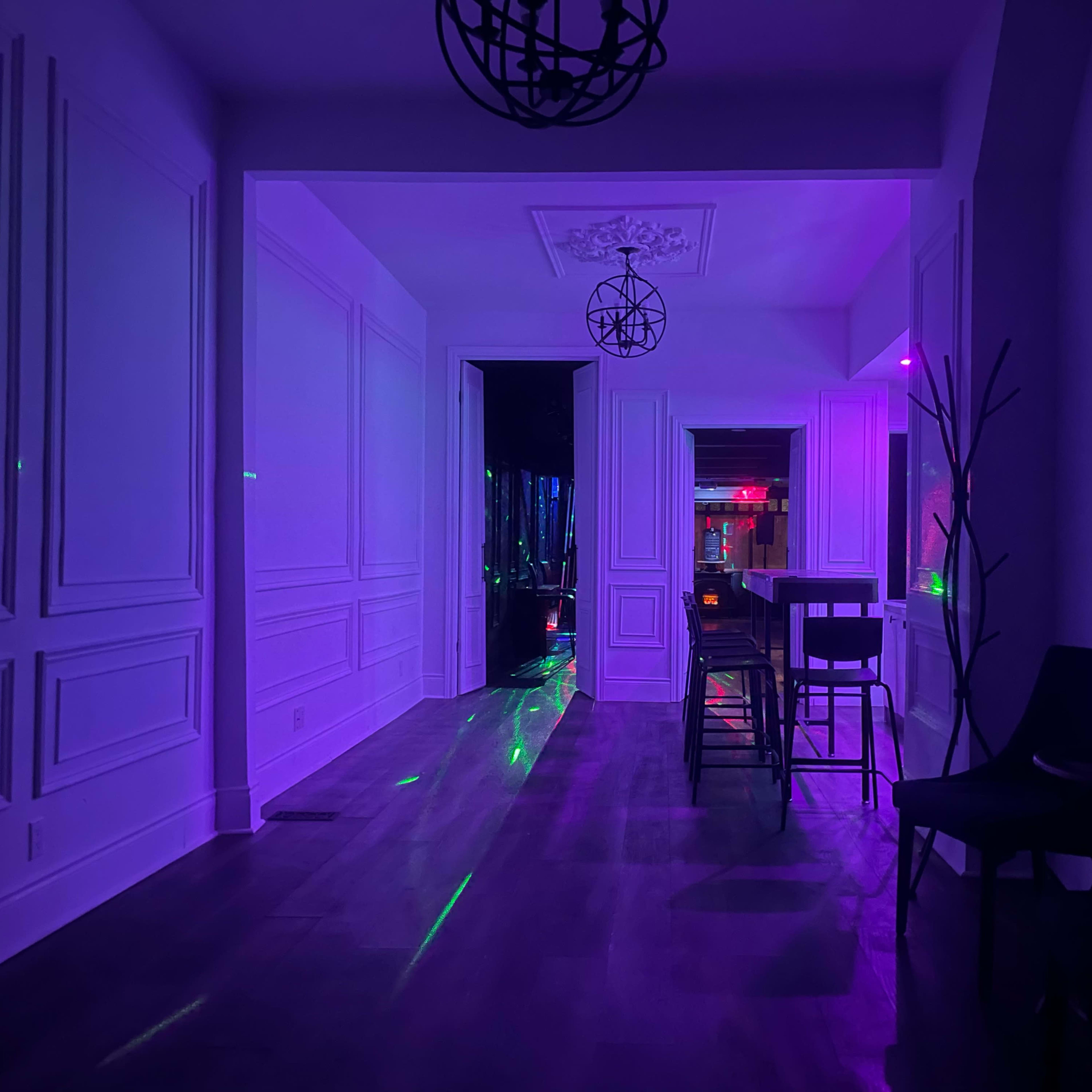 A dimly lit hallway with purple lighting, leading to a room illuminated by colorful lights and featuring bar stools and a wooden table.