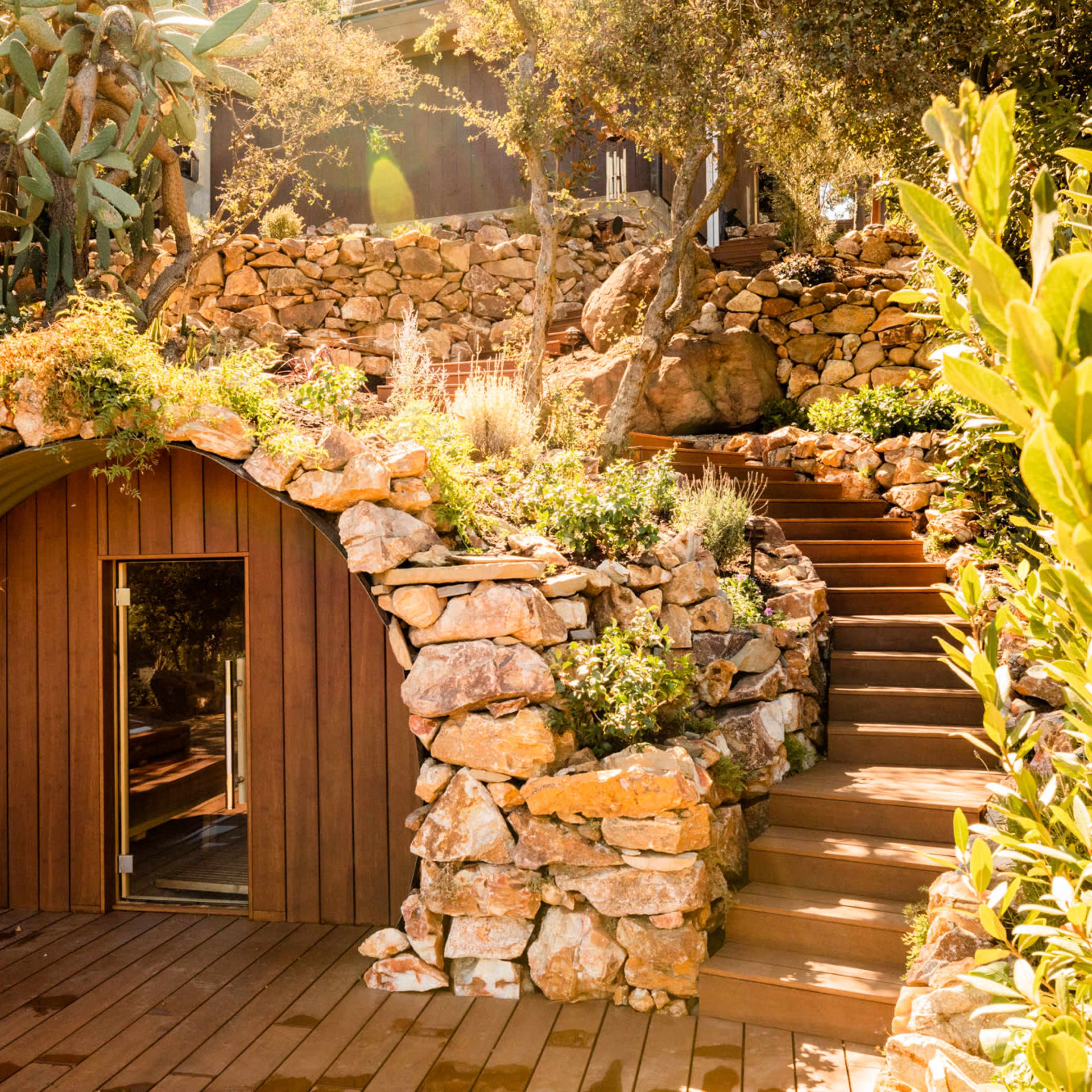 A curved stone structure set into a hillside, surrounded by lush greenery and steps leading up to a wooden deck.