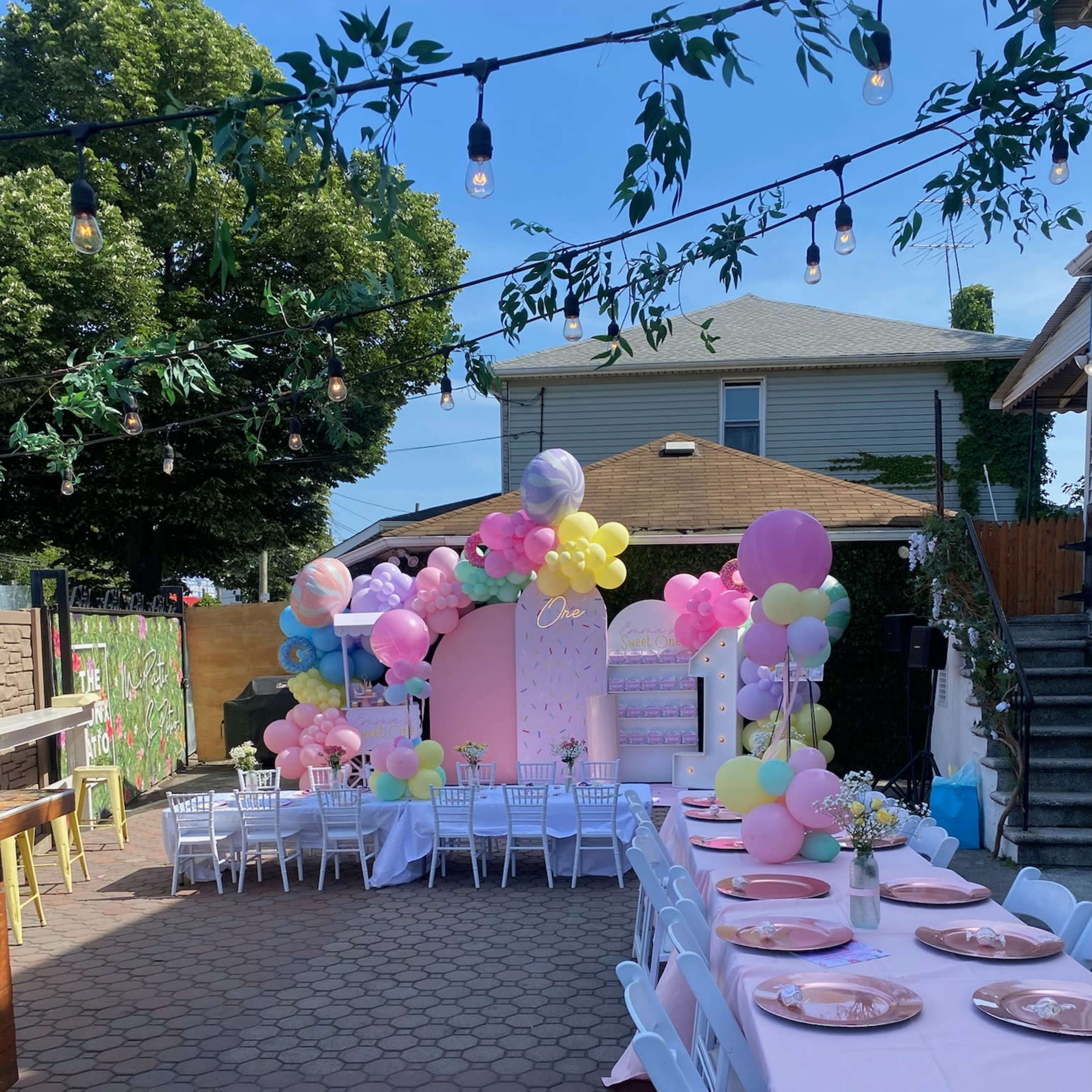 A decorated outdoor space features a long table set with pink plates and chairs, surrounded by colorful balloon arrangements and a festive backdrop.