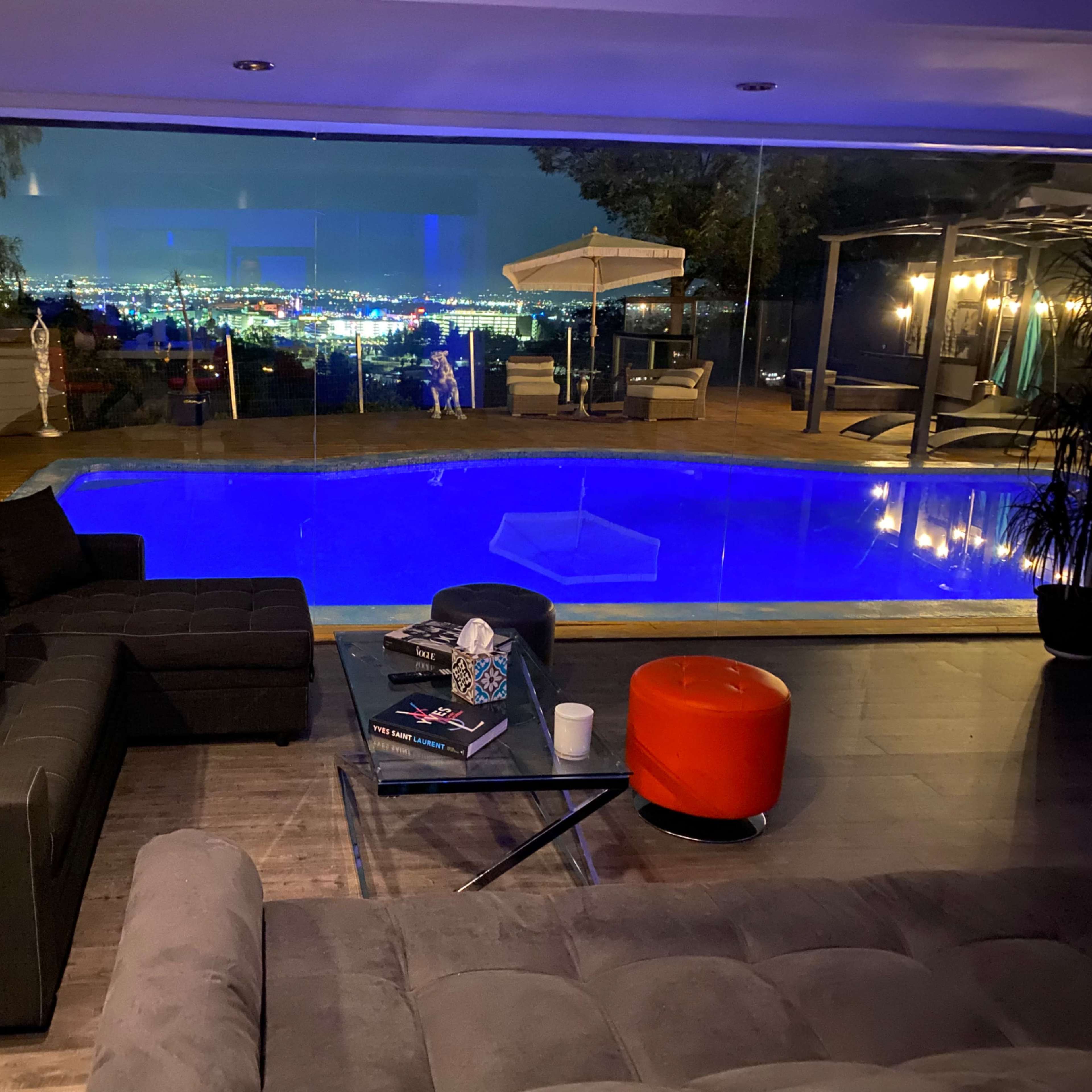 The scene shows a modern living area with a black sectional sofa and an orange ottoman, overlooking a brightly lit outdoor pool and city skyline at night.