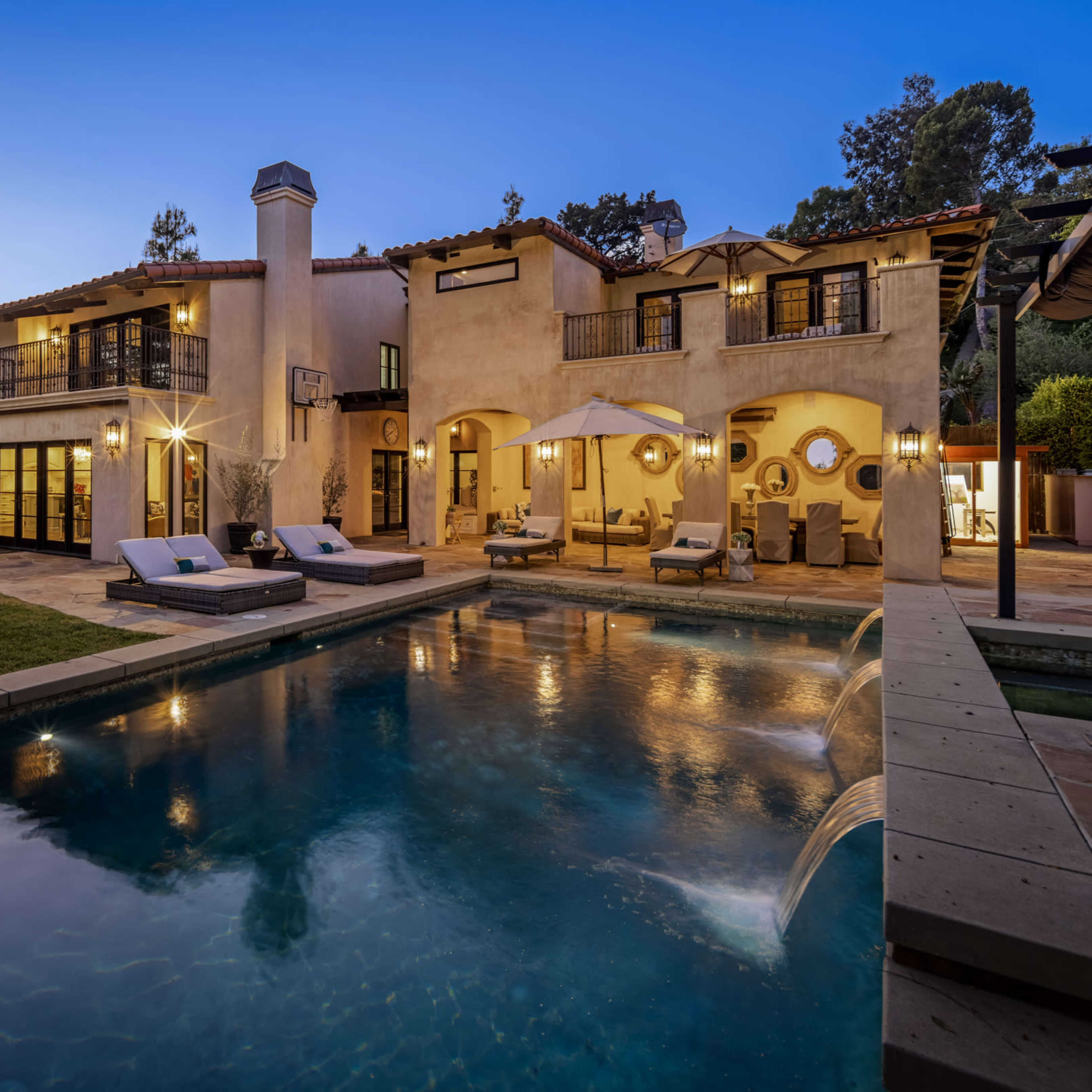 A two-story villa with a pool and outdoor seating areas, surrounded by greenery and illuminated in the evening light.