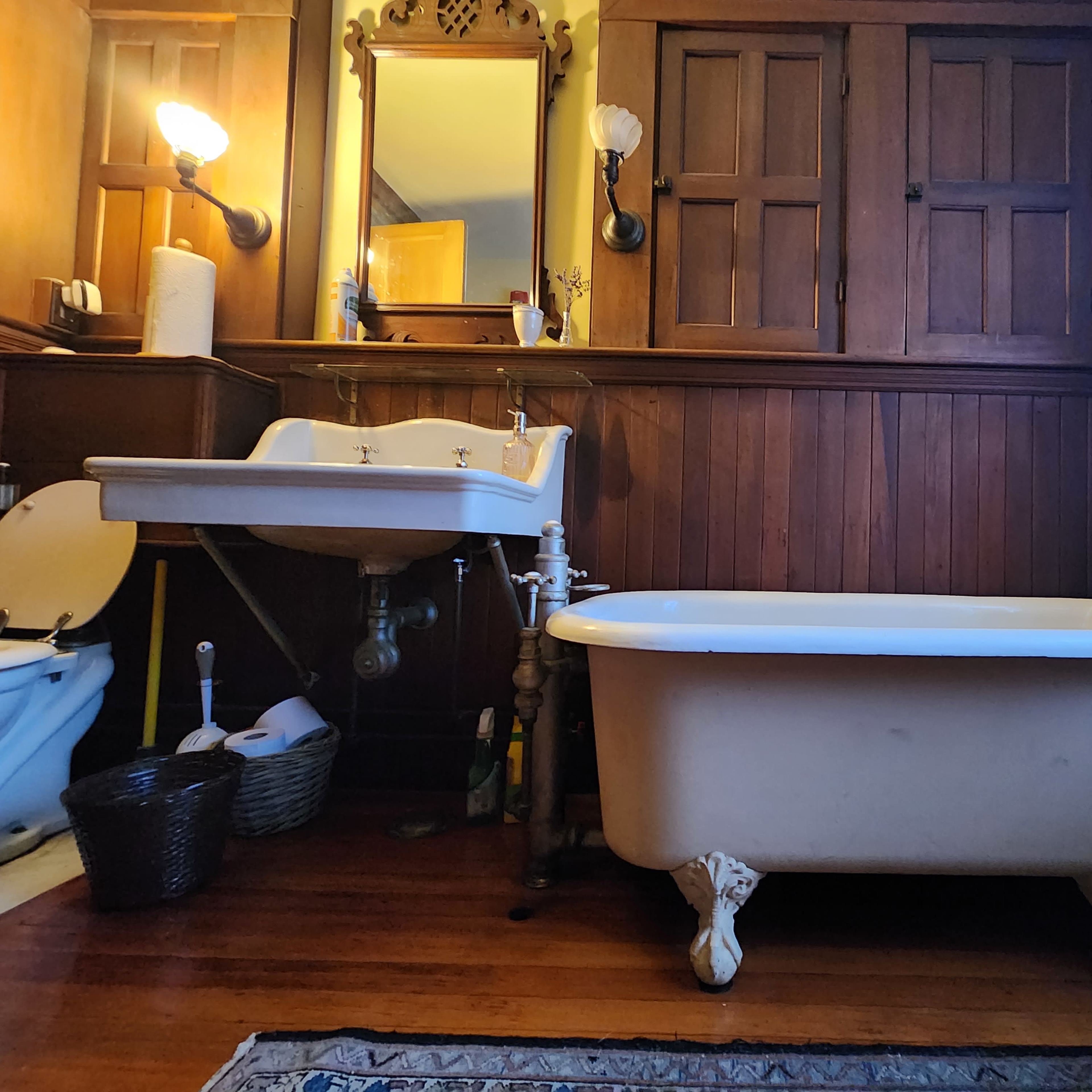 A vintage bathroom with a clawfoot bathtub, a pedestal sink with faucets, a wooden cabinet, and dark wood paneling on the walls.