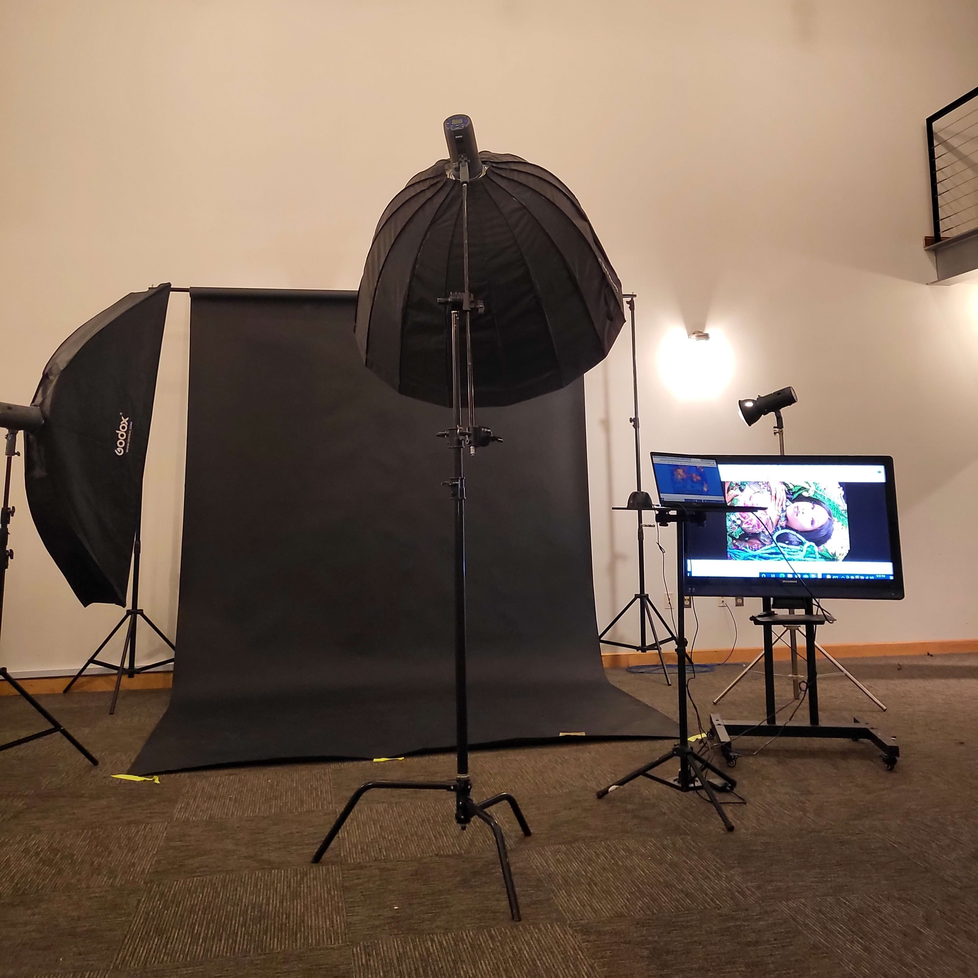 A photography studio setup, featuring softbox lights, a dark backdrop, and a computer monitor displaying an image.