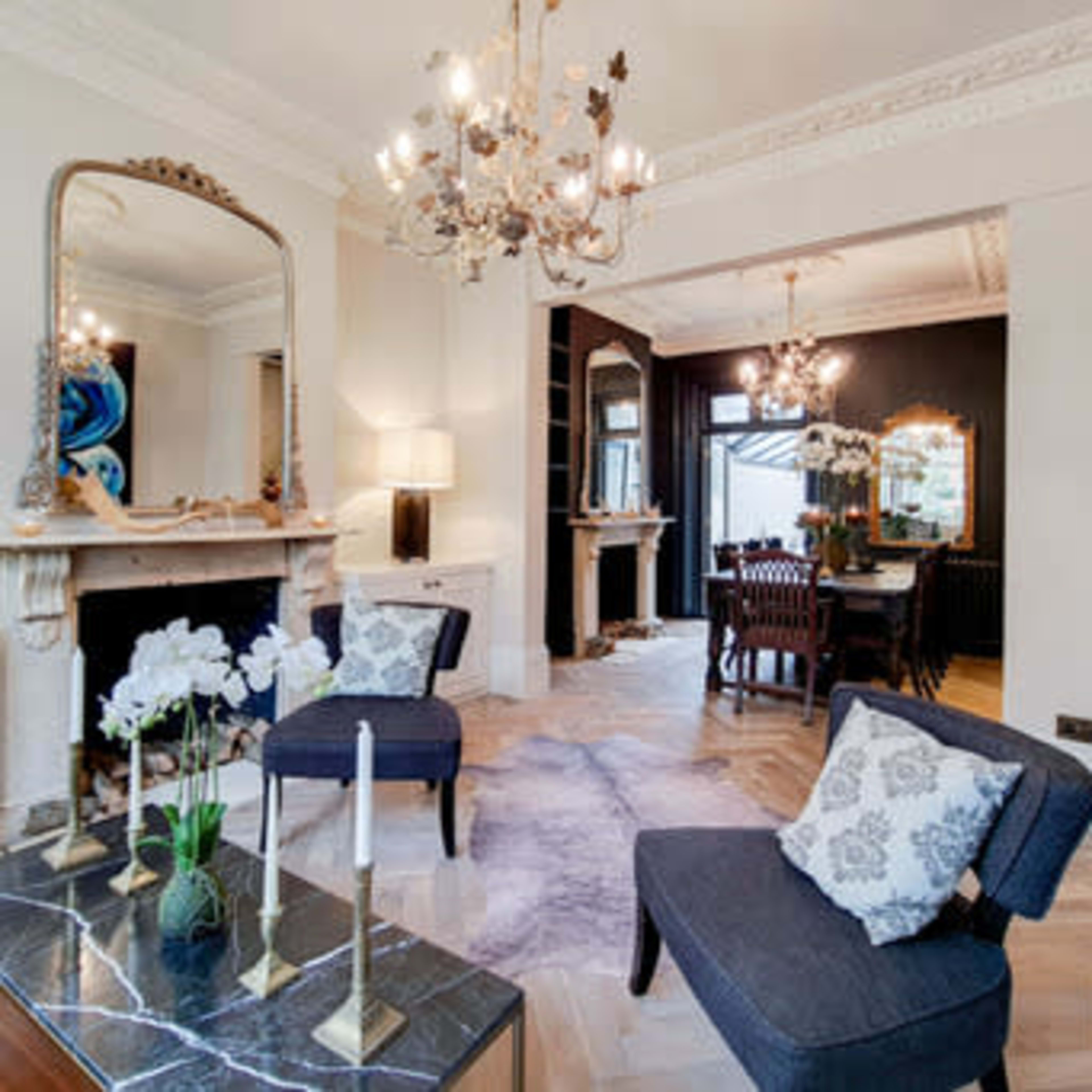 A stylish living space featuring a marble coffee table, two dark armchairs, a large mirror, and a chandelier, with a dining area visible in the background.
