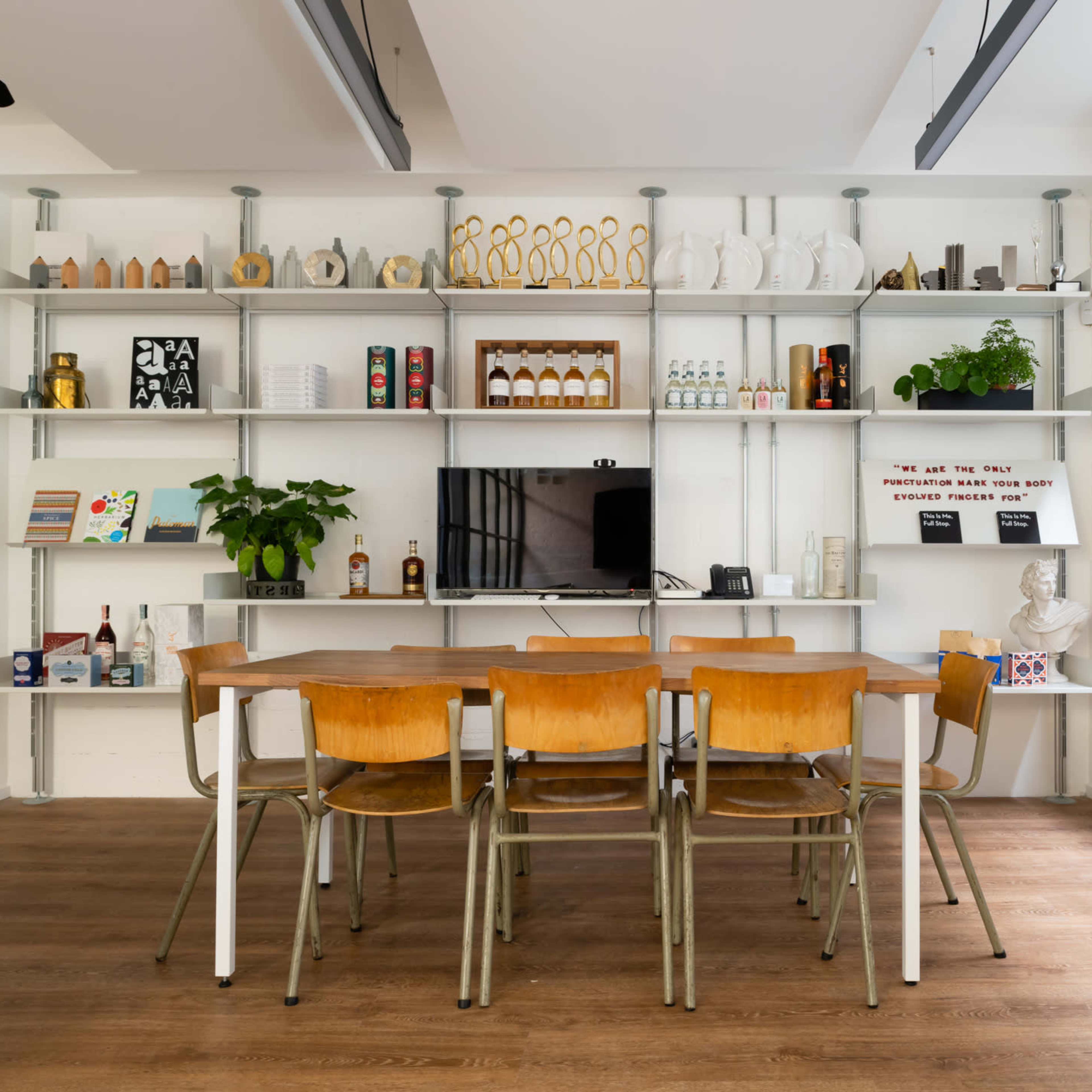 A modern workspace with a wooden table surrounded by chairs, and shelves displaying various decorative items and bottles.