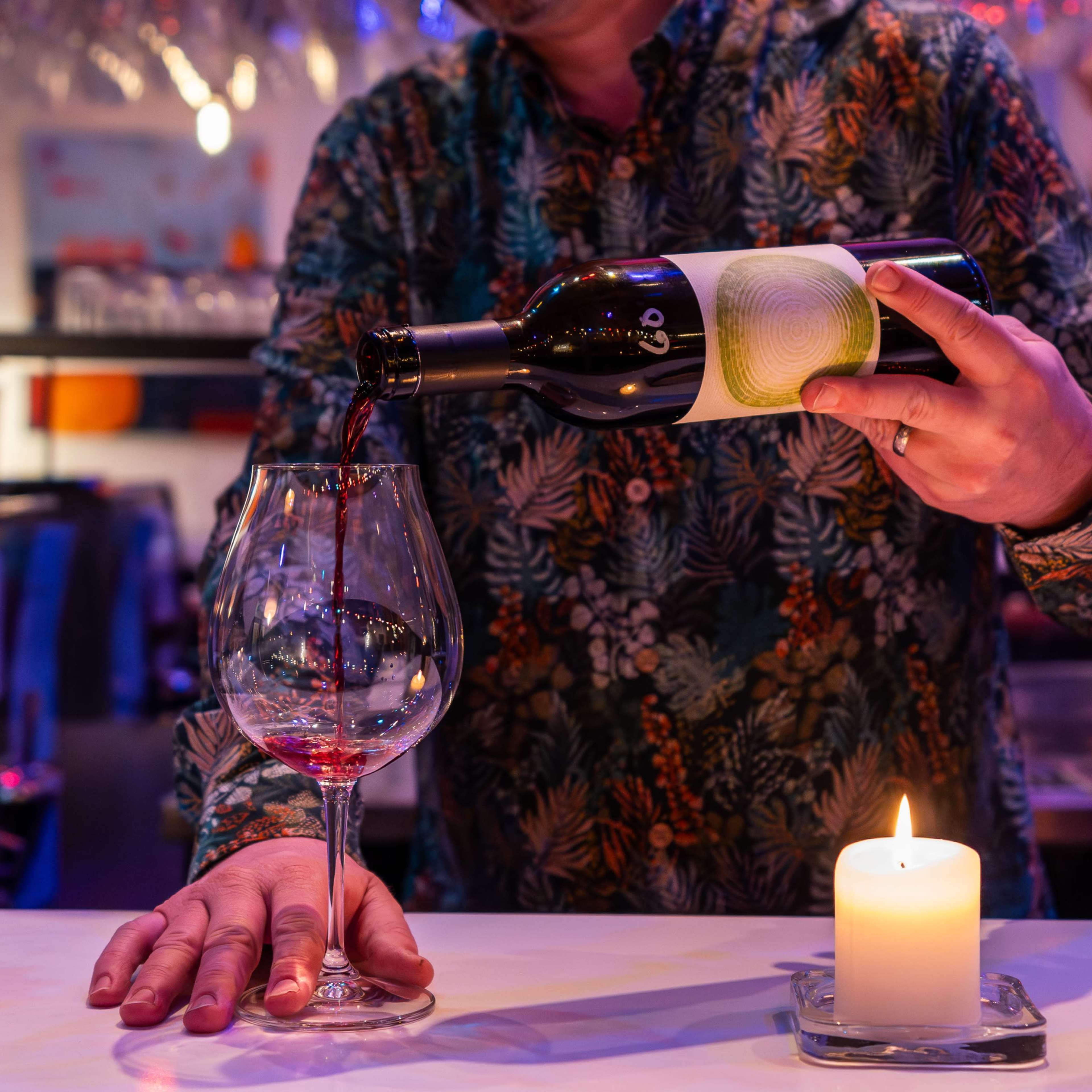 A person in a patterned shirt pours red wine from a bottle into a glass at a bar, while a candle burns on the counter.
