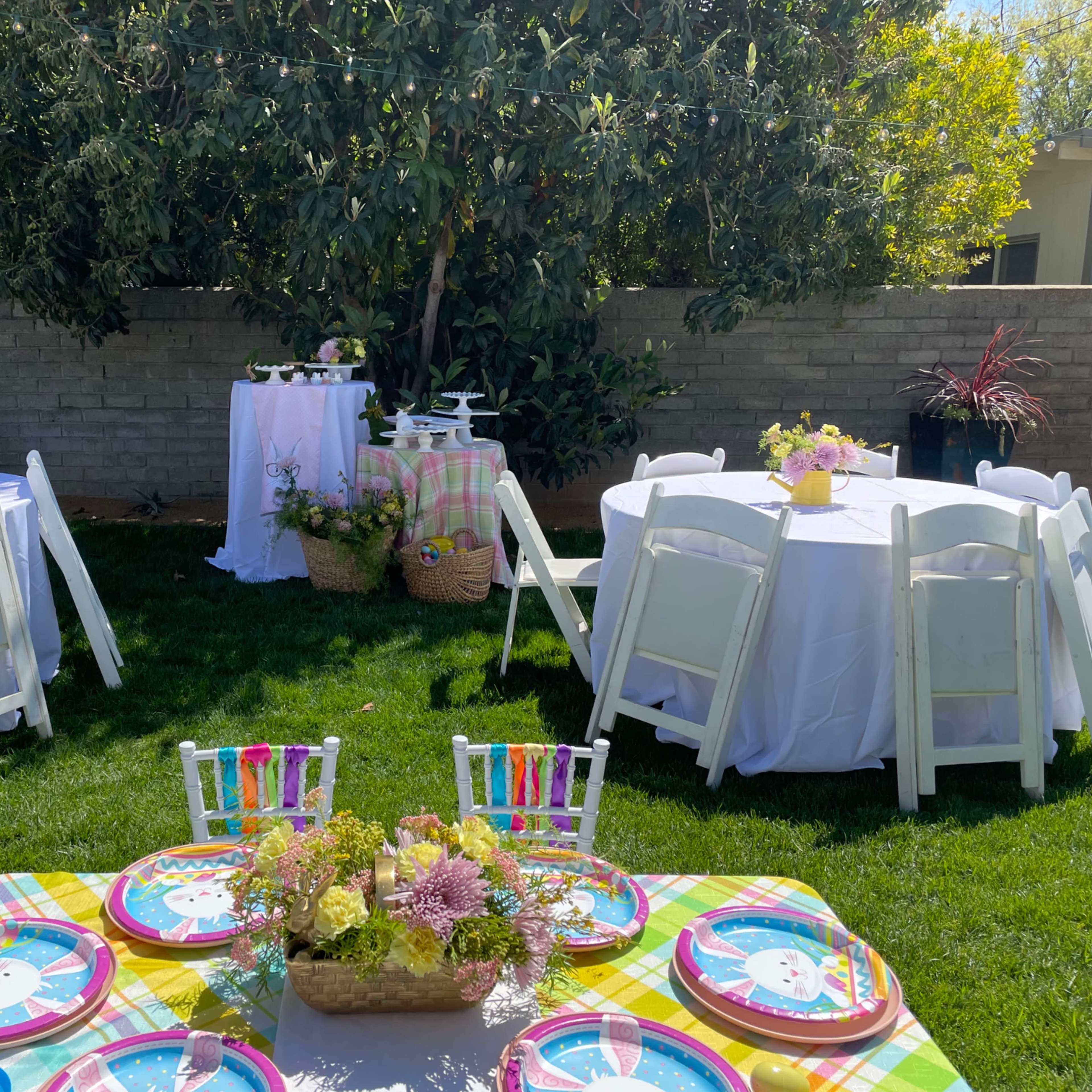 The scene features an outdoor party setup with round tables covered in white tablecloths, colorful place settings, and floral centerpieces on a grassy lawn.