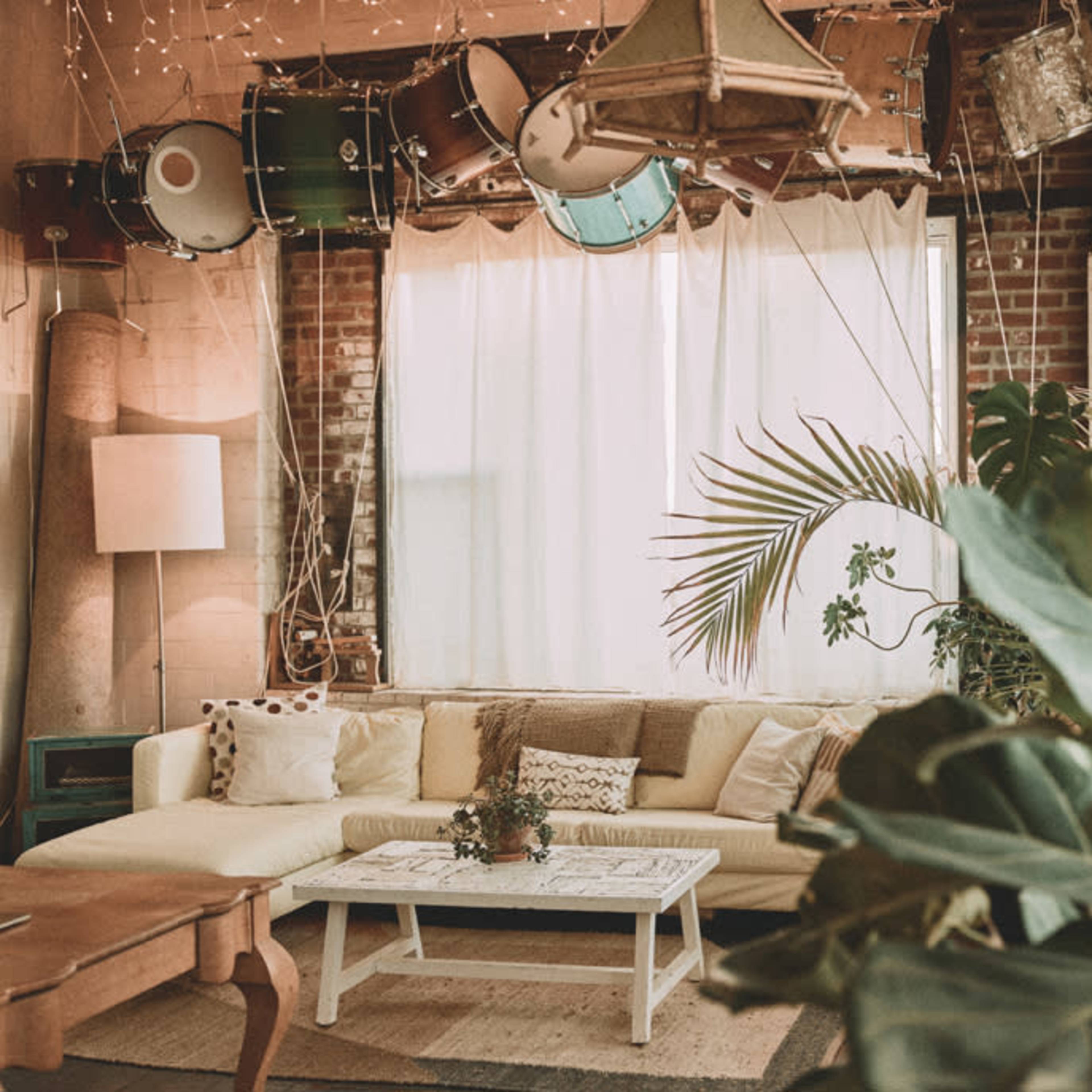A cozy living room with a white sofa, a coffee table, and plants, featuring a backdrop of large windows and hanging drums.