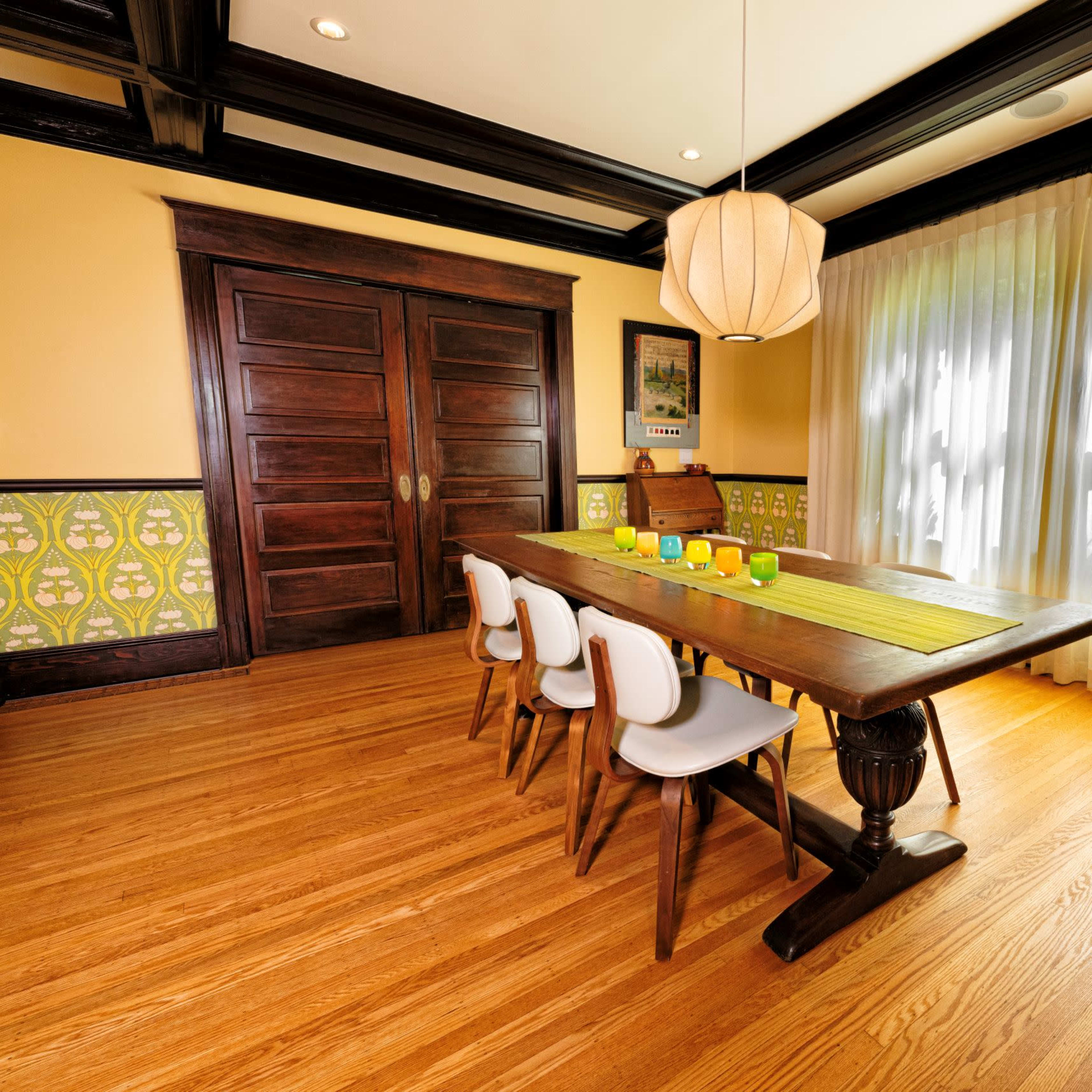 A wooden dining table with chairs is set in a well-lit room featuring yellow walls, a large window covered with sheer curtains, and decorative paneling.