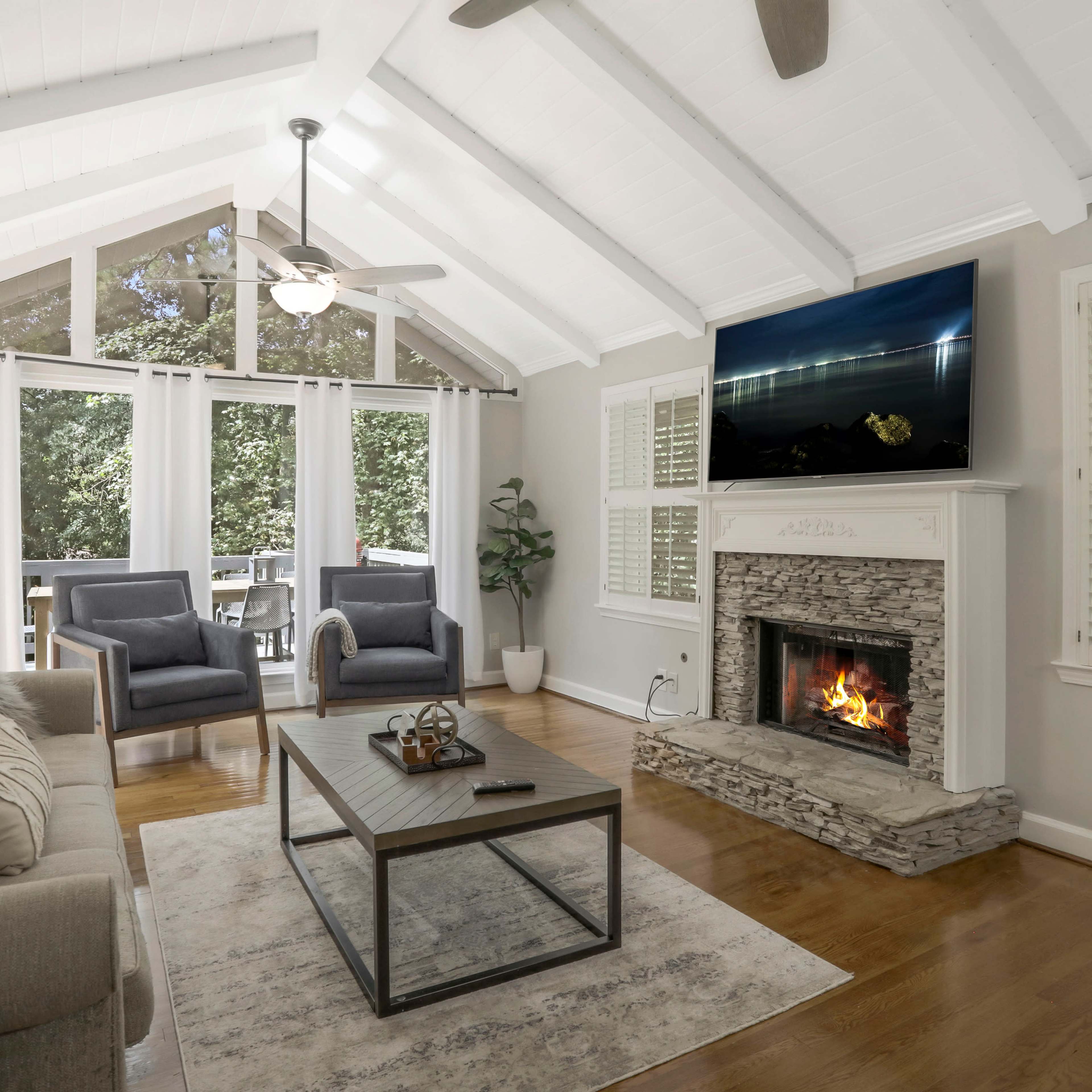 A bright living room features a stone fireplace, a large television mounted above it, comfortable seating, and large windows overlooking a green view.