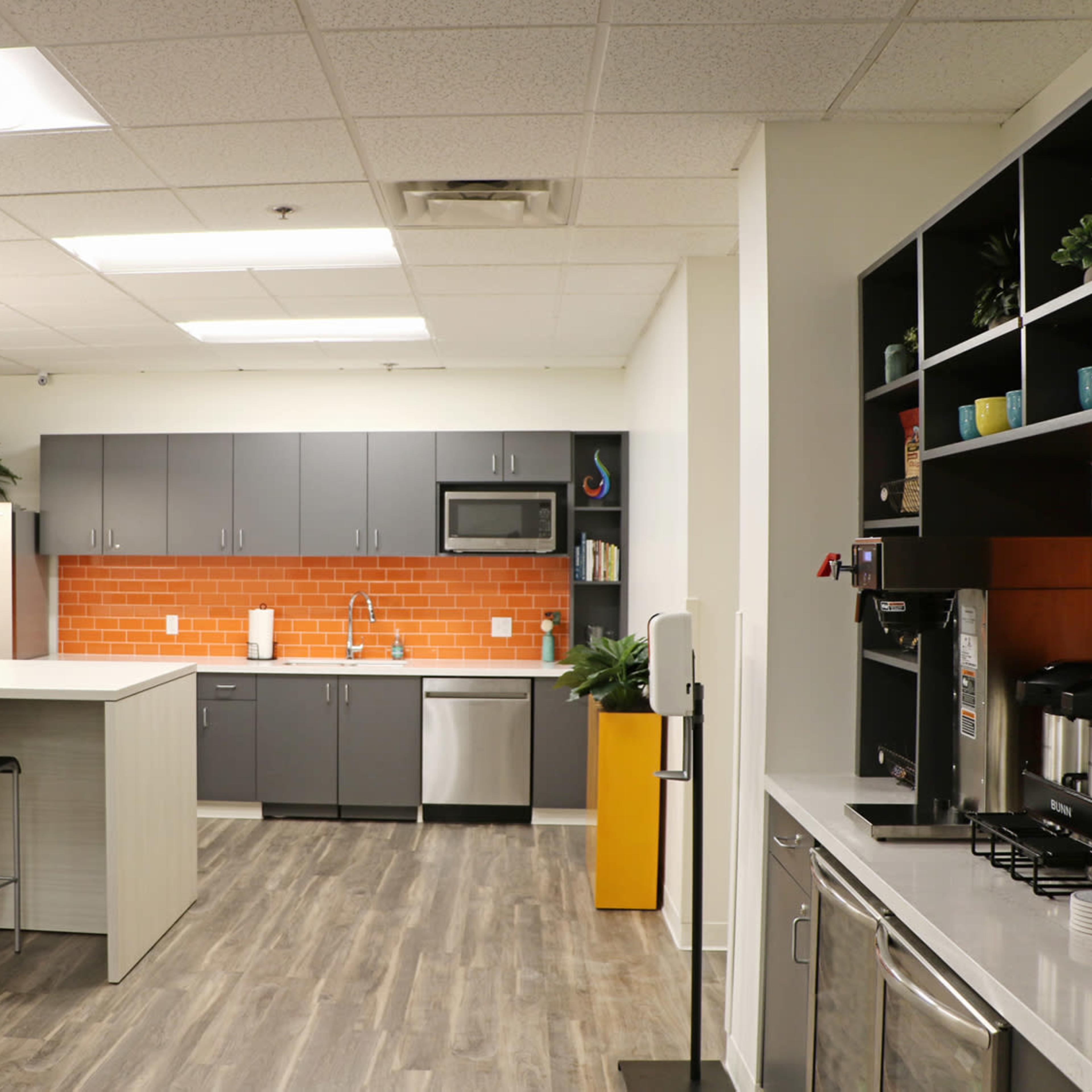 A modern kitchen features gray cabinetry, stainless steel appliances, and a central island with bar stools, accented by an orange tiled backsplash.