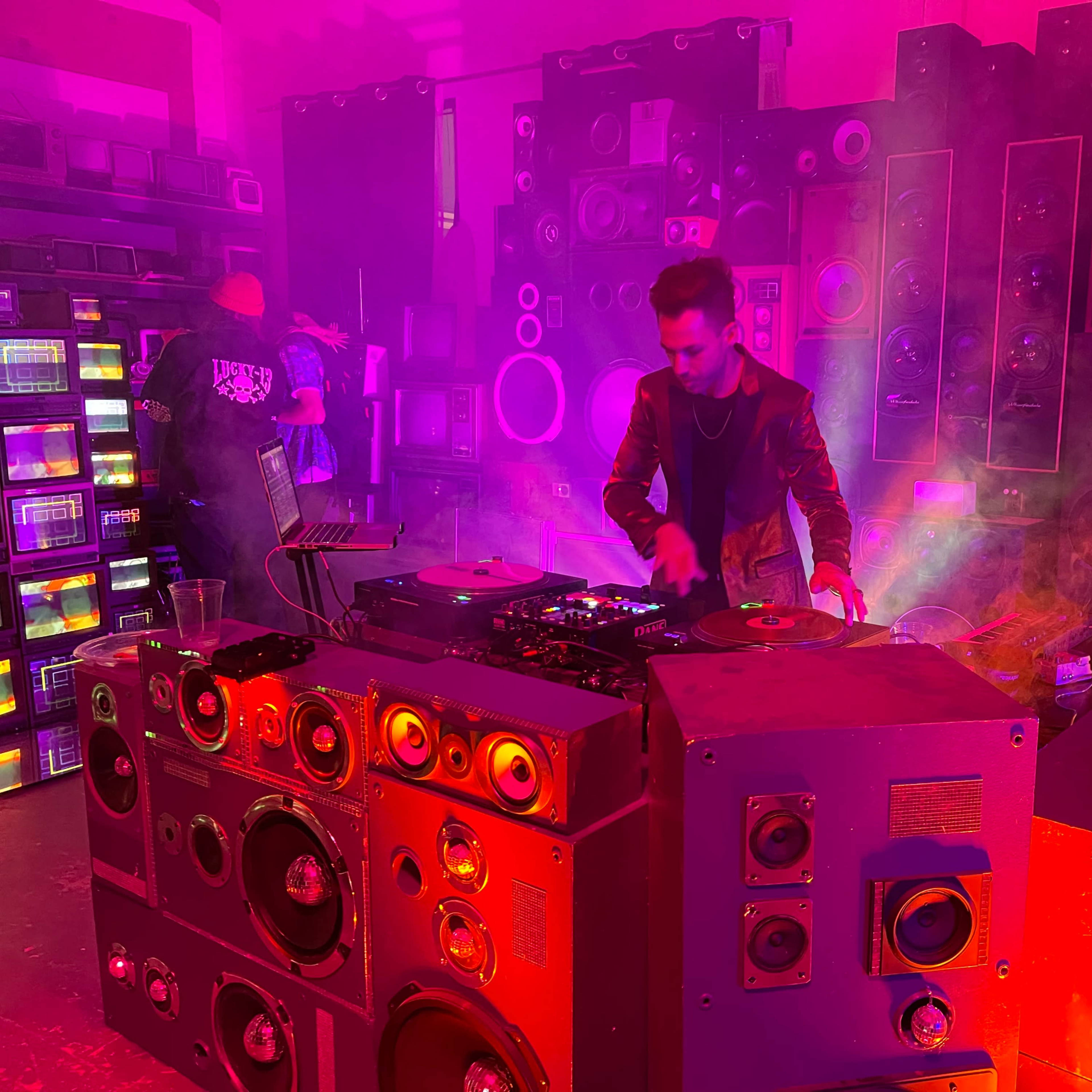 A DJ performs in a room filled with colorful, stacked speakers while vibrant lights illuminate the atmosphere.