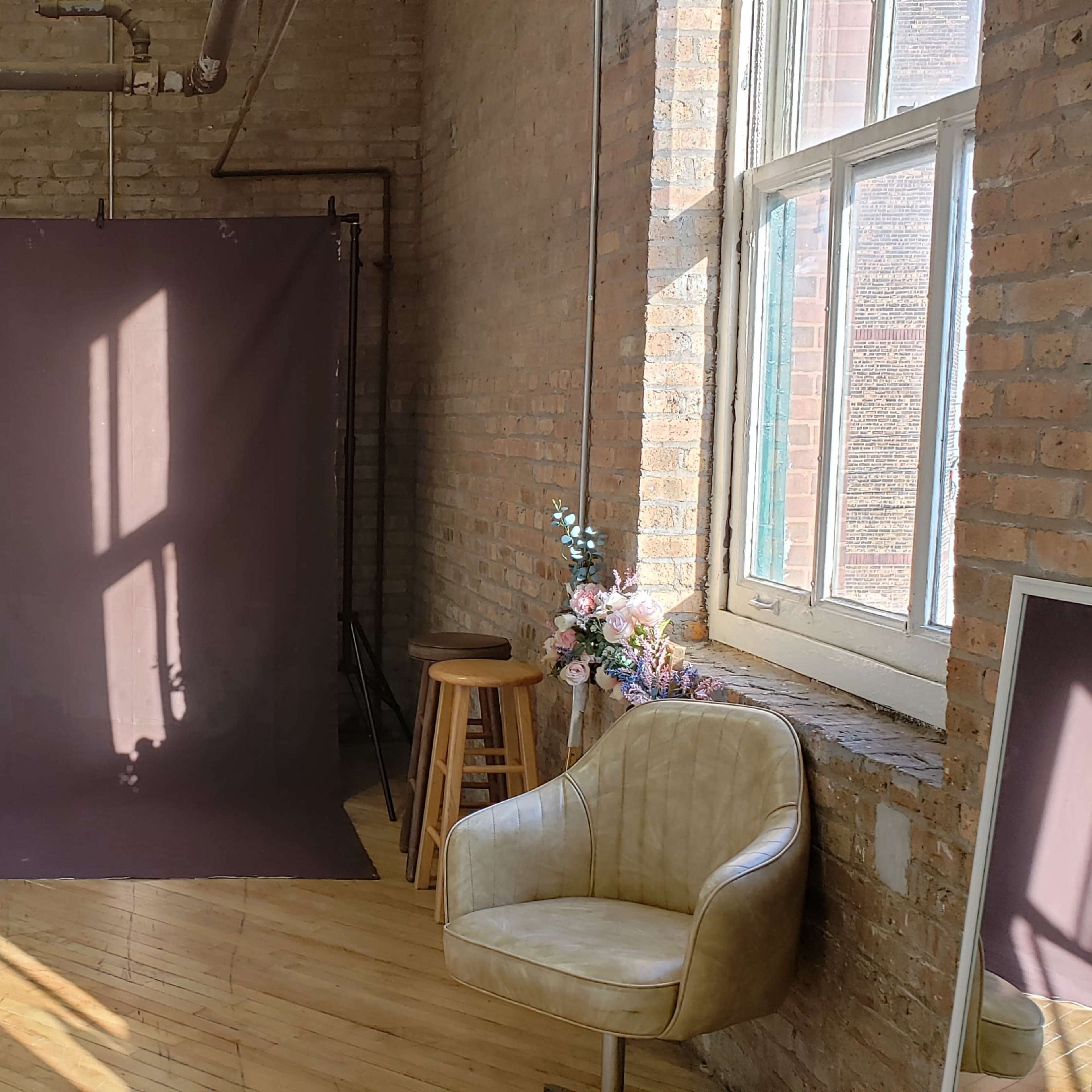 A spacious room features a brown backdrop, wooden flooring, a chair, and shelves with plants against a brick wall.