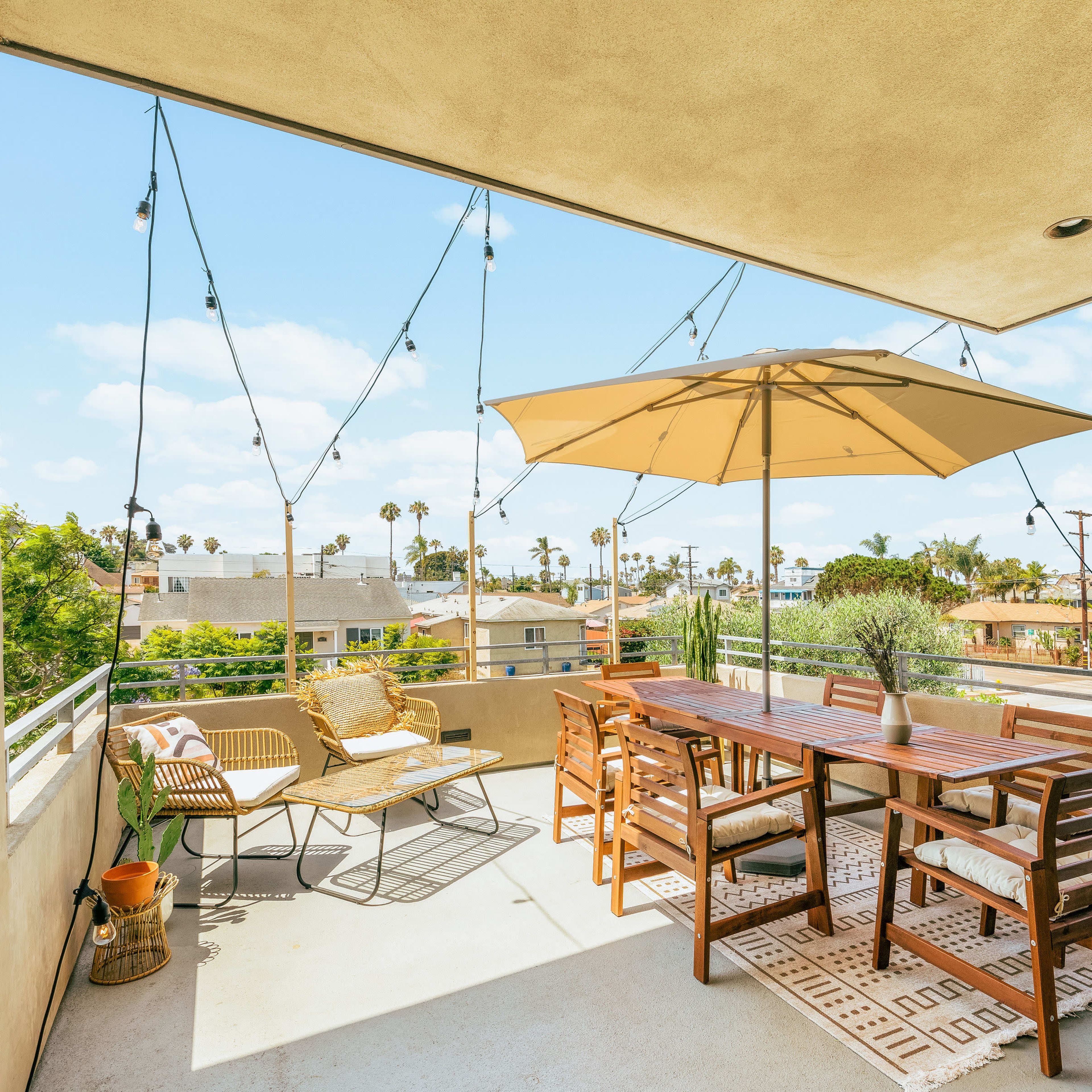 A spacious outdoor terrace features a dining table with an umbrella, string lights overhead, and seating arrangements with palm trees in the background.