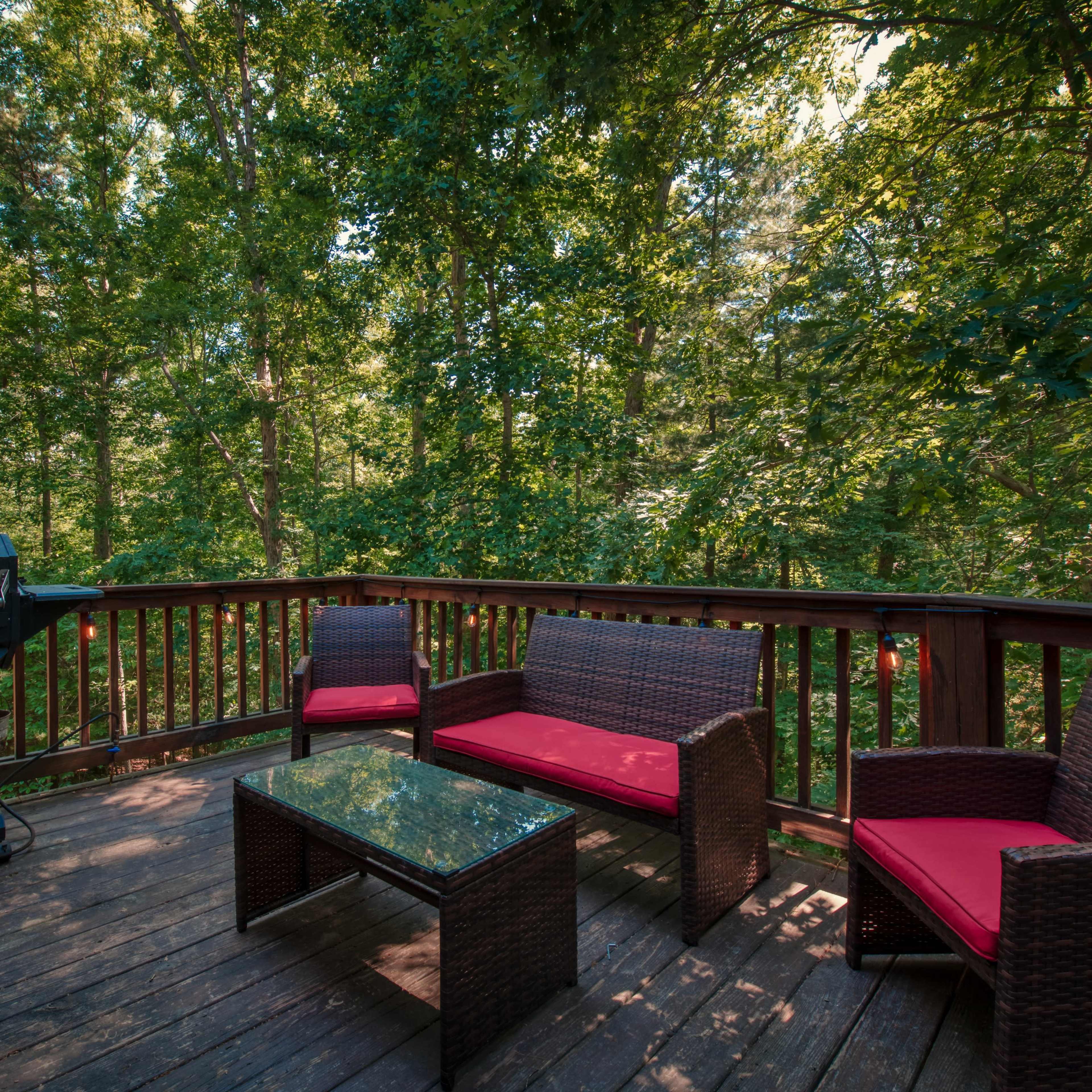 A wooden deck features a grill and outdoor furniture surrounded by dense greenery.