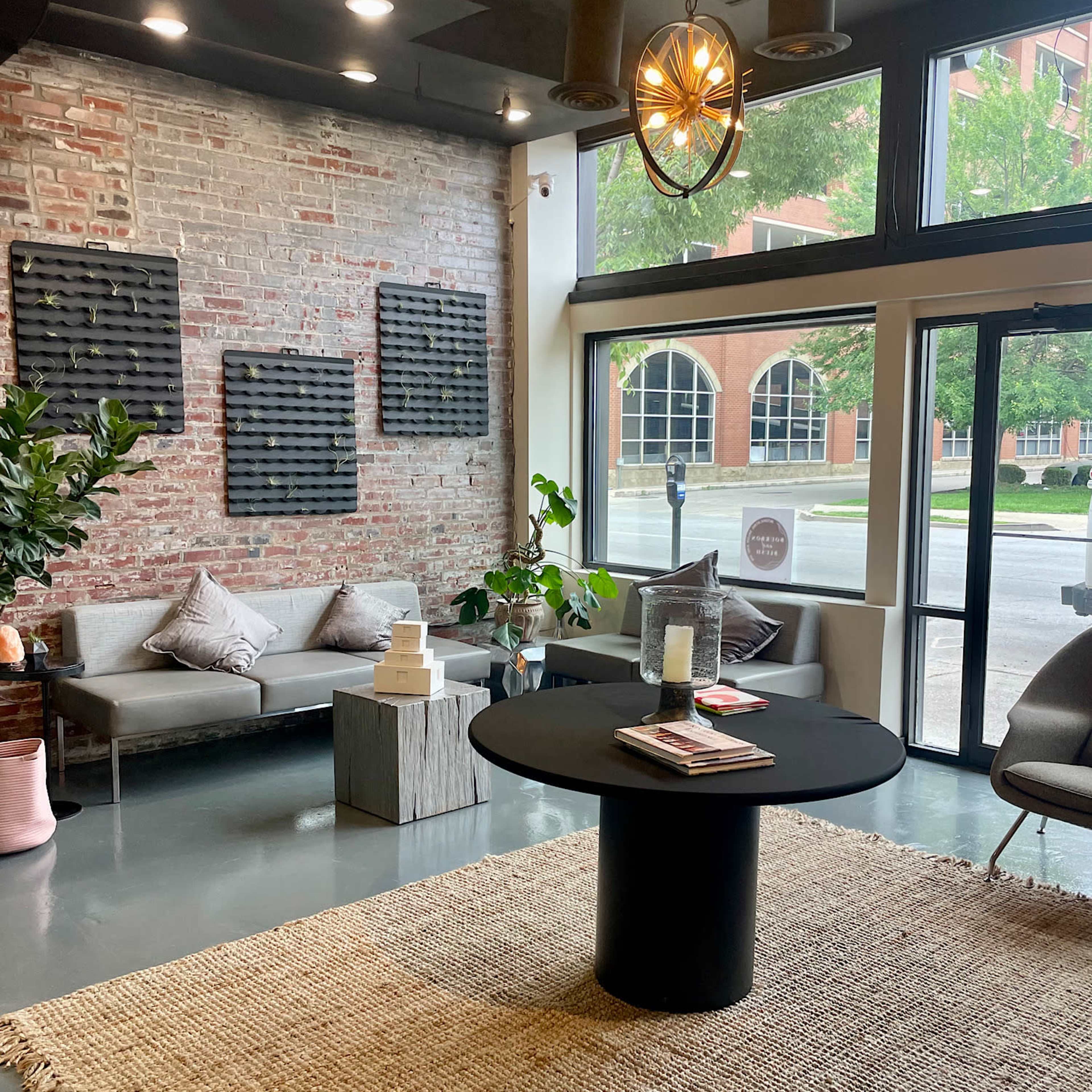 A modern indoor space featuring a seating area with a gray sofa, a round black table, and large windows facing a street, complemented by a woven rug and plants.