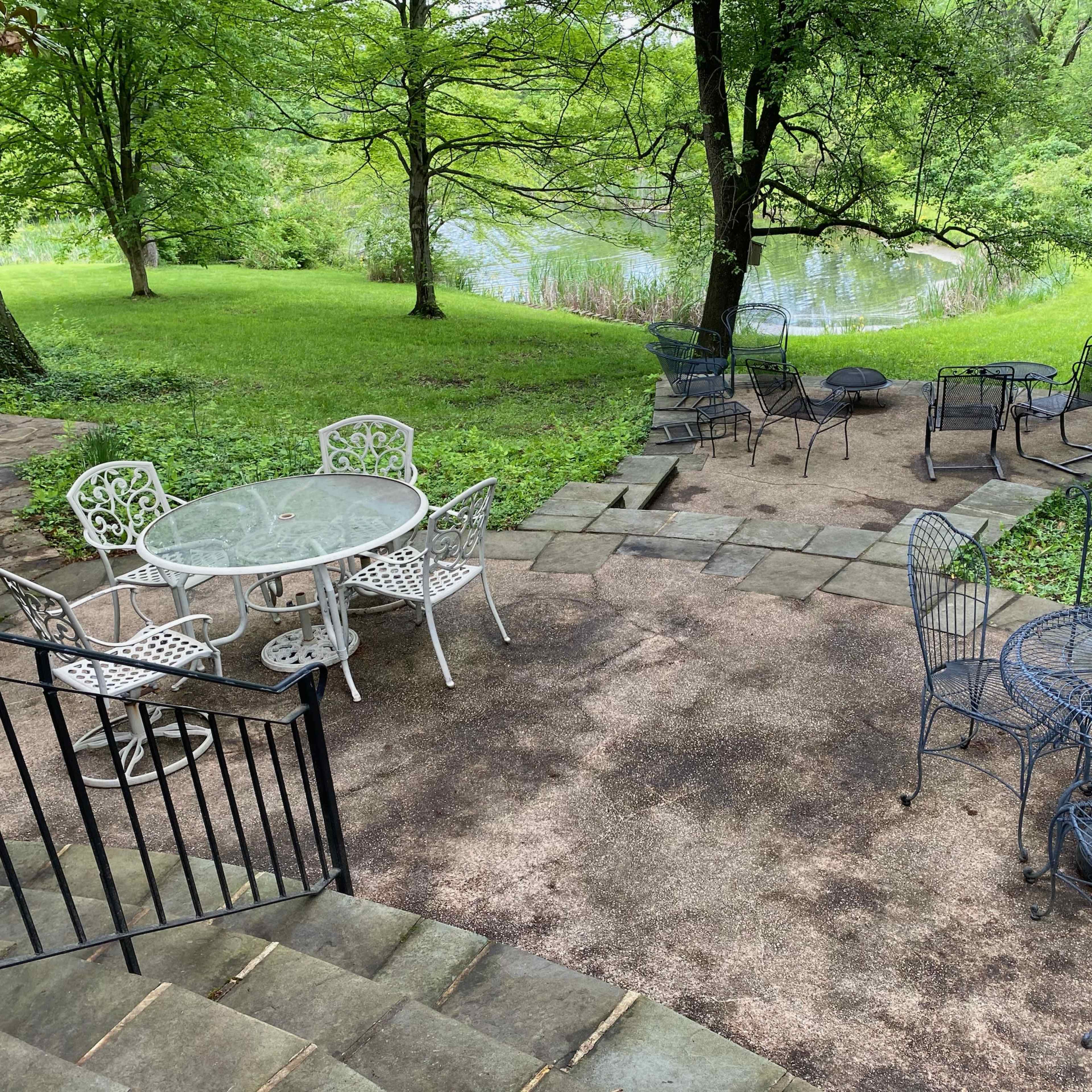 A stone patio features several outdoor tables and chairs arranged near a grassy area and a pond.