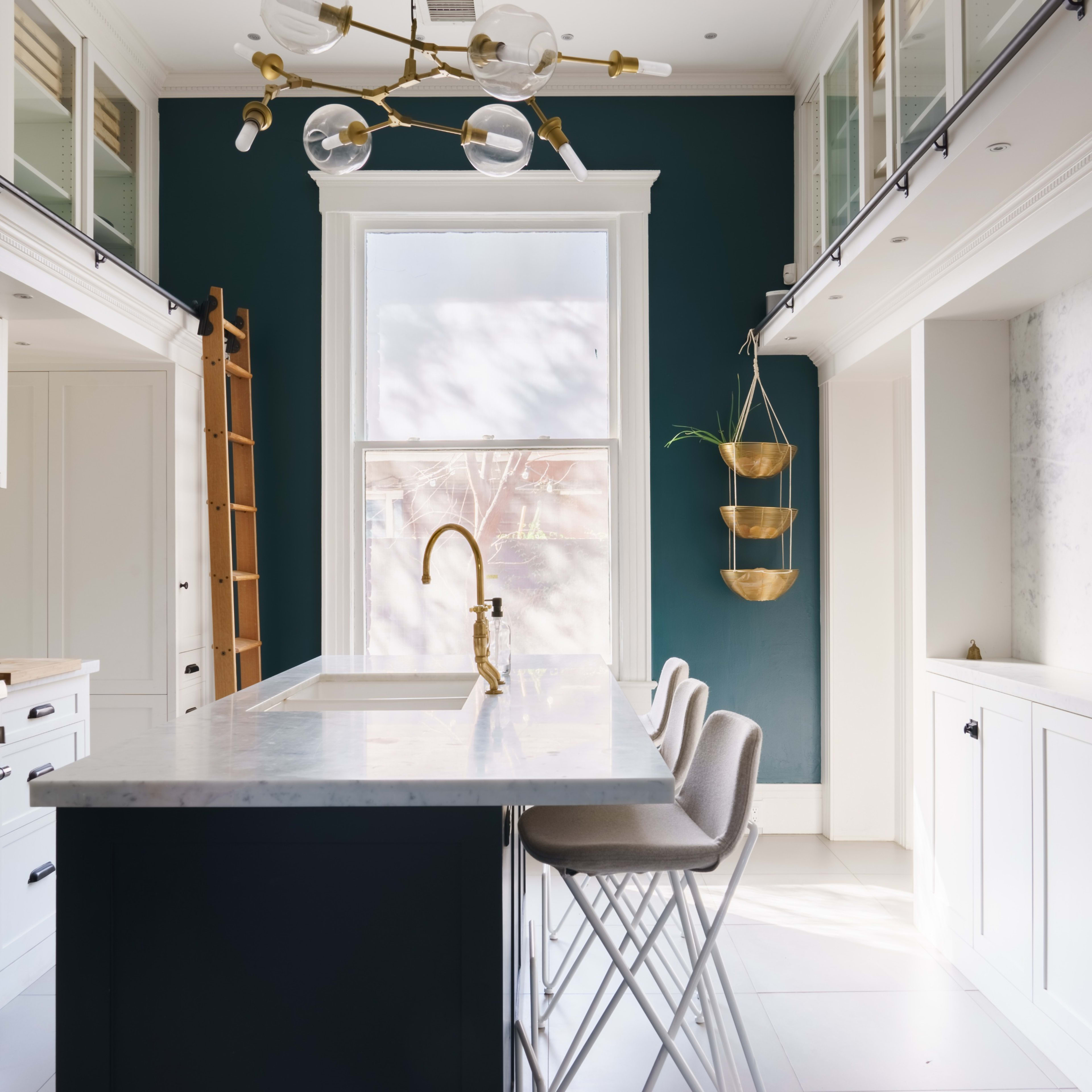 A modern kitchen with a marble island, white cabinetry, a dark blue accent wall, and stylish lighting fixtures.