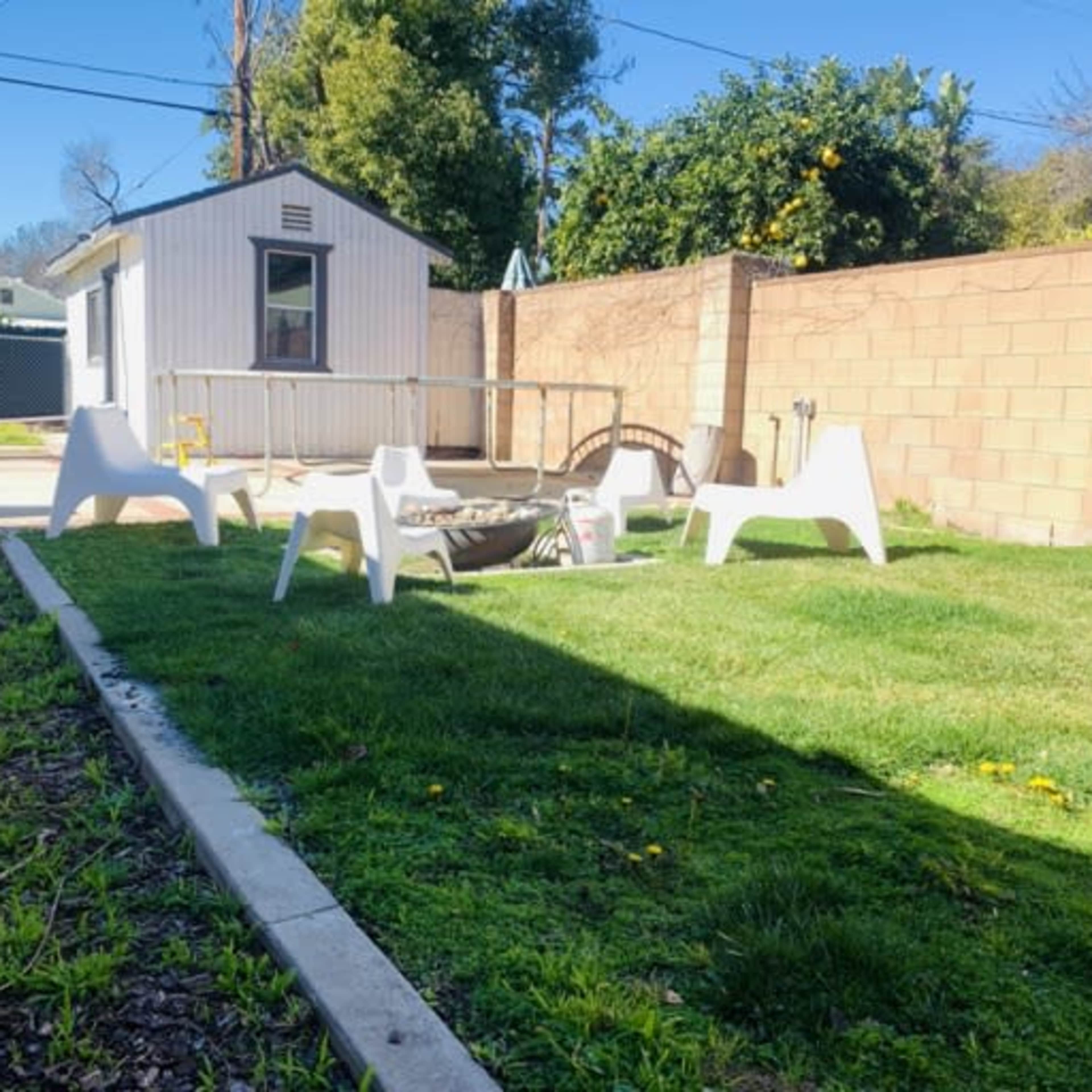 A backyard with a green lawn, white plastic chairs, a circular table, and a small building in the background.