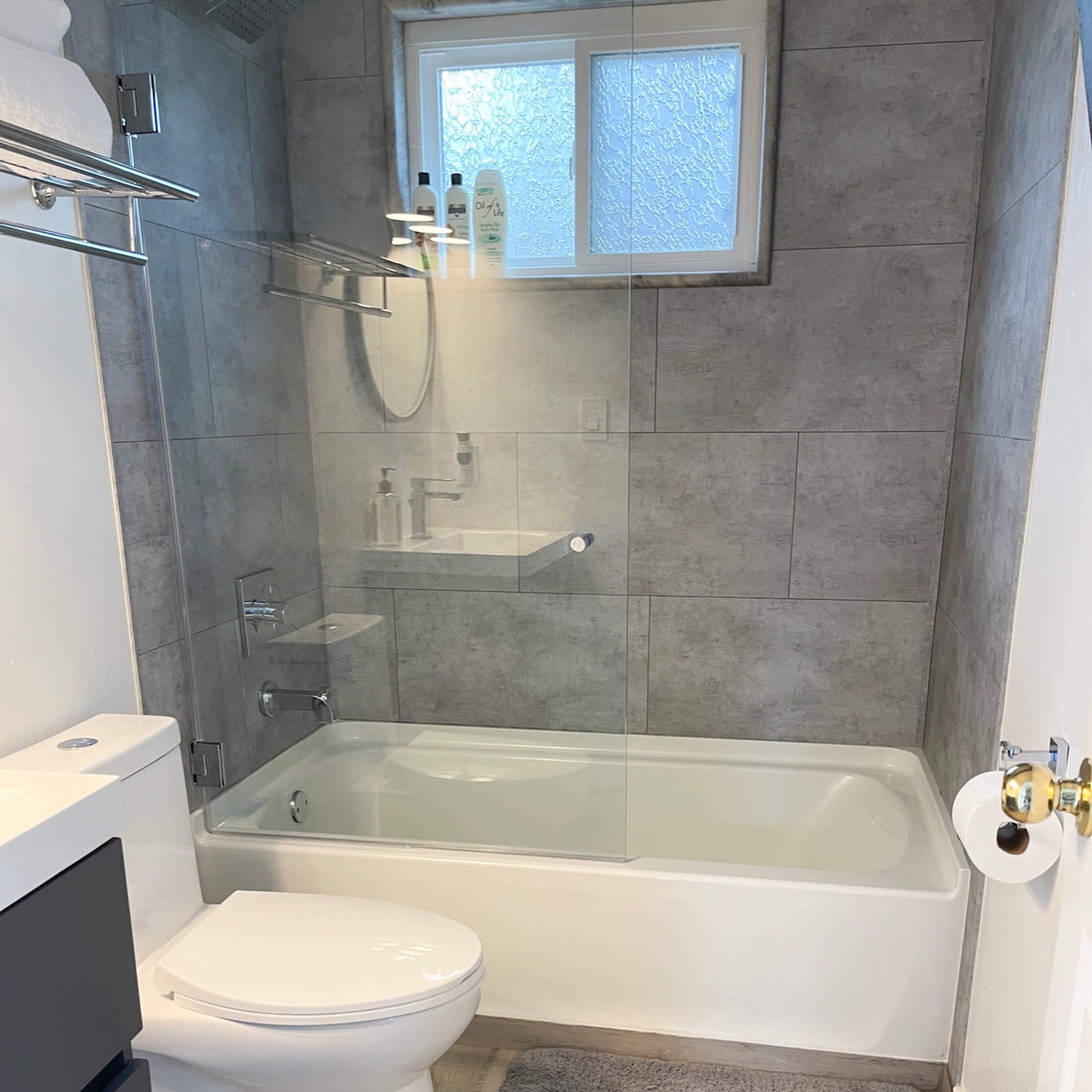 A modern bathroom featuring a combined shower and bathtub, a toilet, and light-colored tile walls with a gray bath mat on the floor.