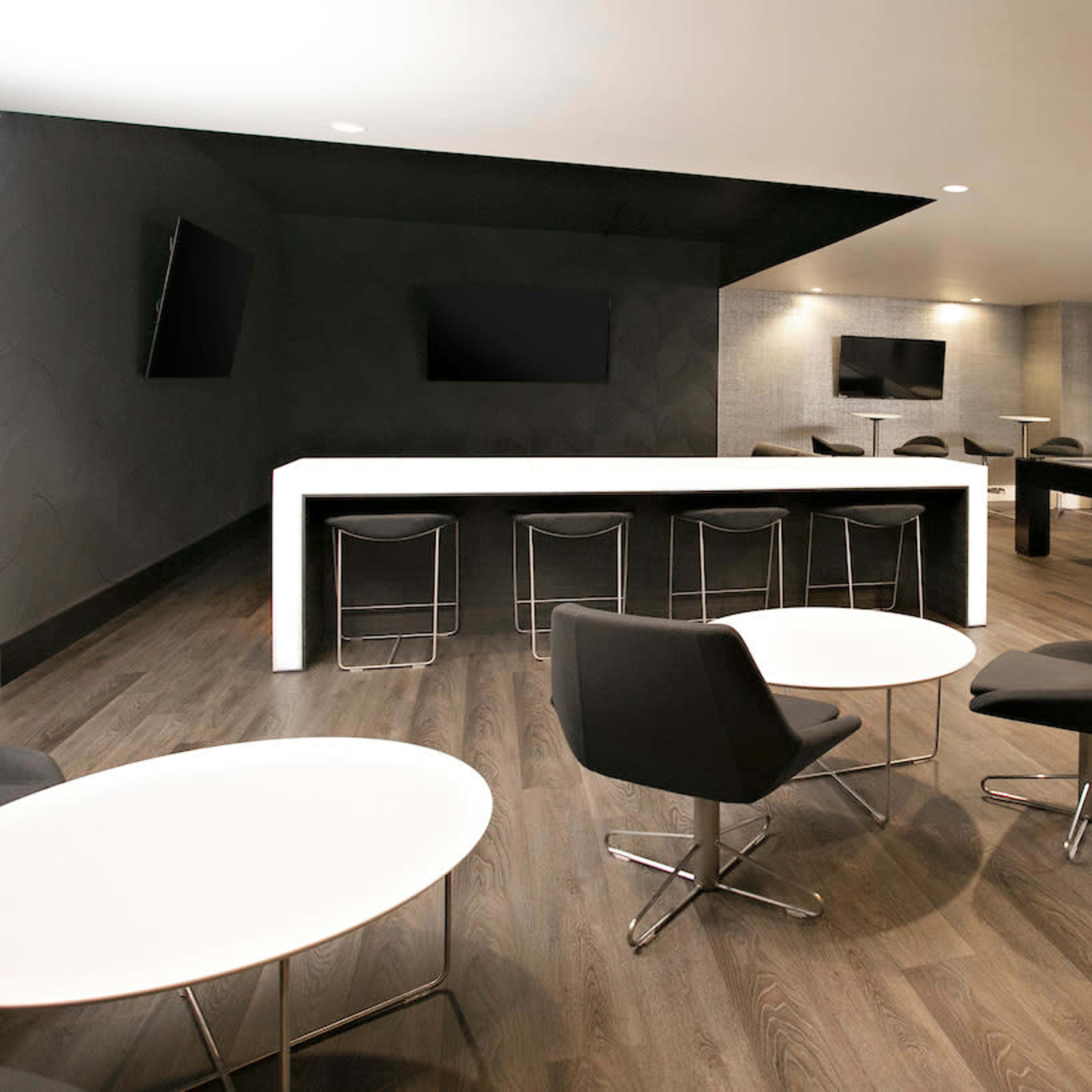A modern lounge area featuring a bar, several round tables, and a billiards table against a sleek, dark wall.