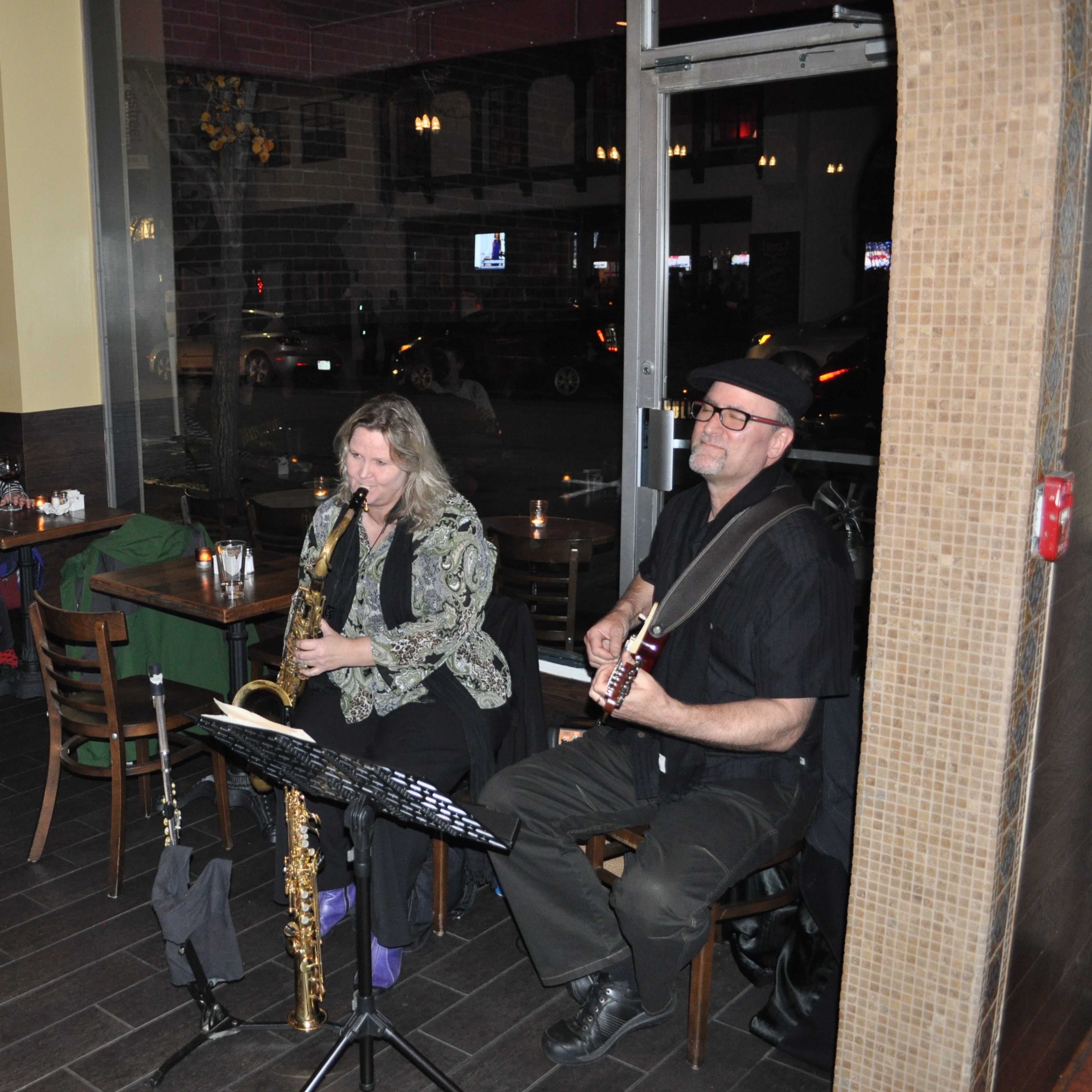 A saxophonist and a guitarist perform in a dimly lit restaurant, while patrons sit at tables nearby enjoying their meals.