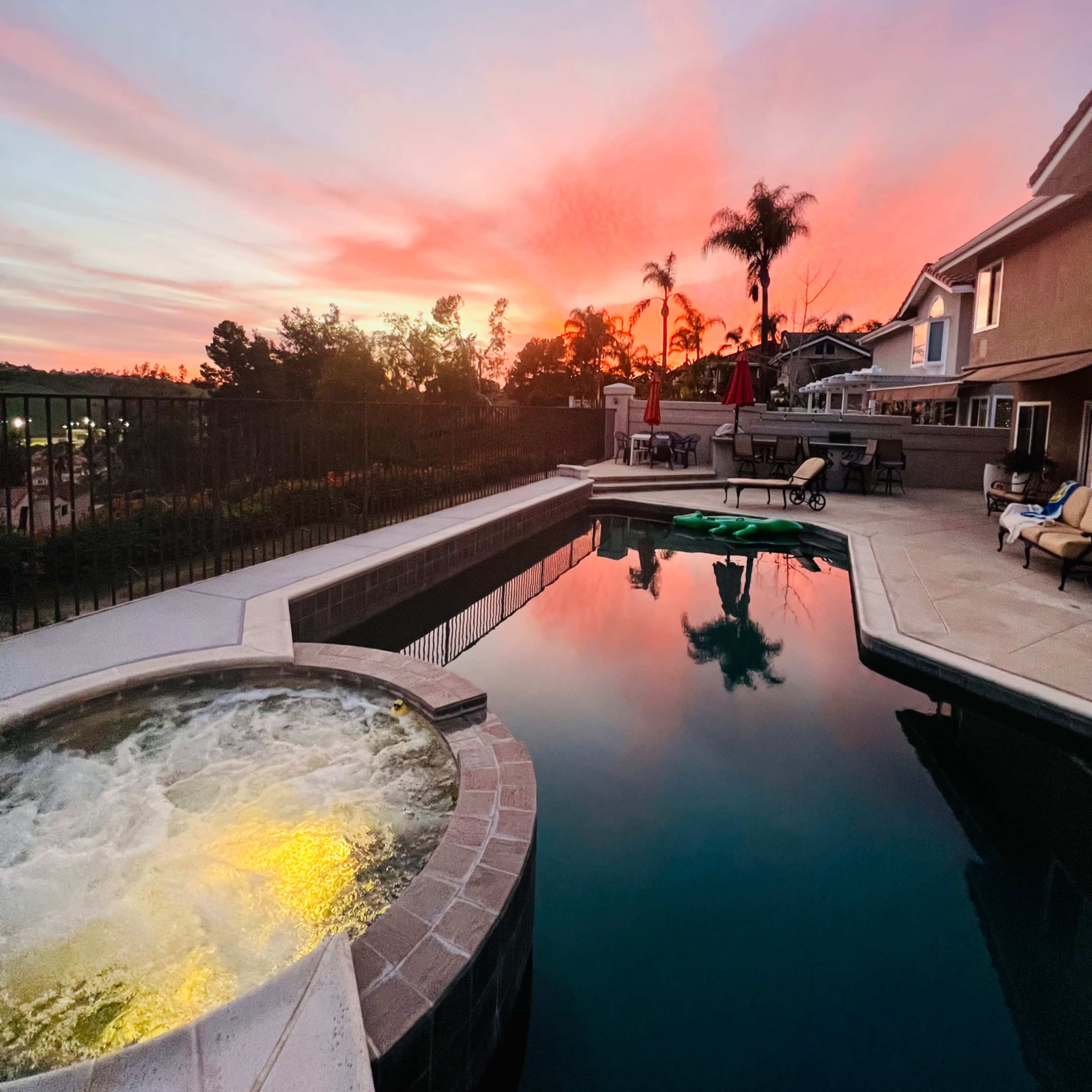 A swimming pool and hot tub are shown at sunset, with reflections of colorful clouds and palm trees in the water.